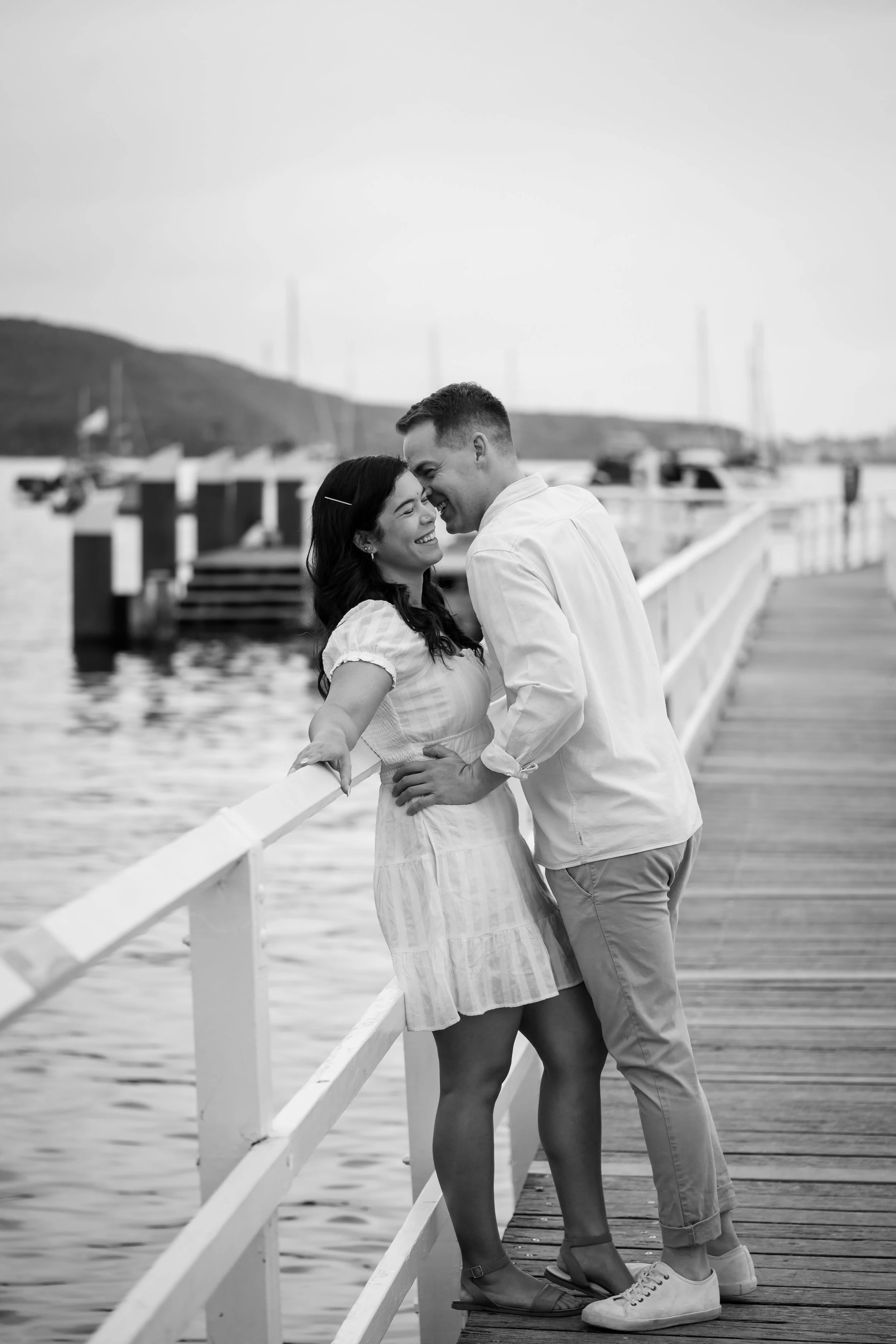 A black and white photo of a couple standing on a dock by the water, smiling and leaning close to each other.