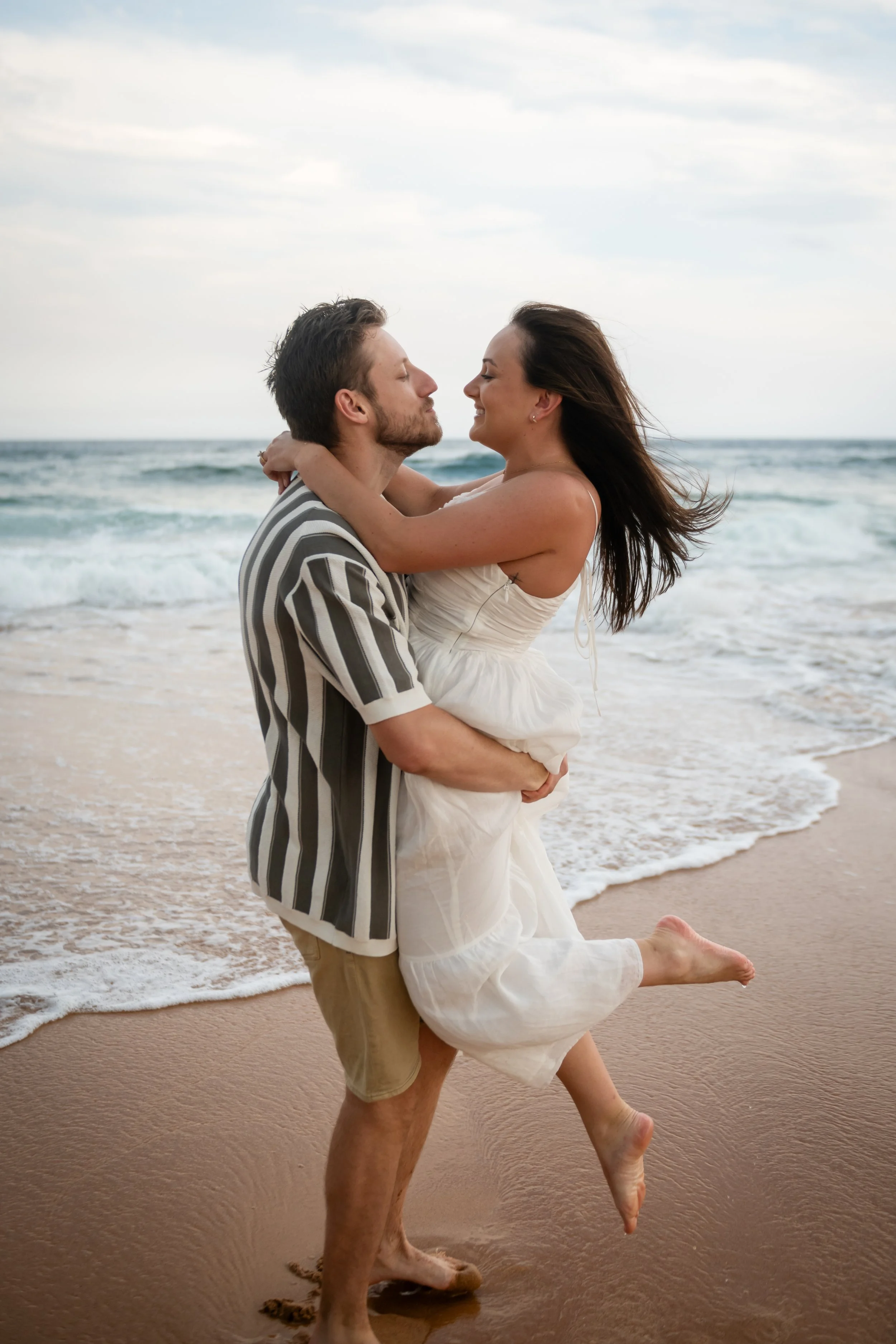 A happy couple is embracing on the beach, with the man lifting the woman in his arms. They are smiling and looking into each other's eyes, with the ocean and cloudy sky in the background.