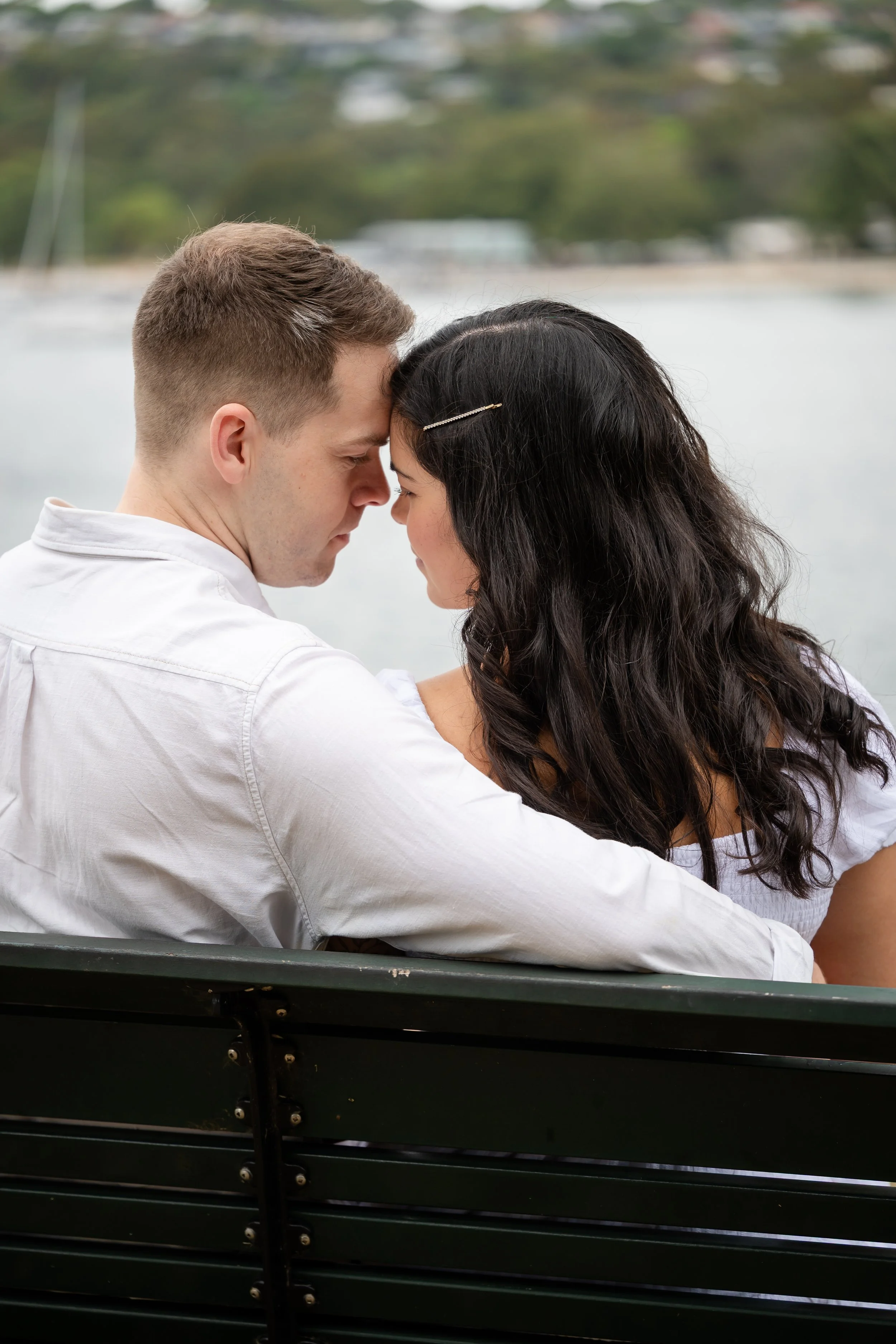 A young couple sitting closely on a park bench with their foreheads touching, overlooking a body of water with trees in the background.