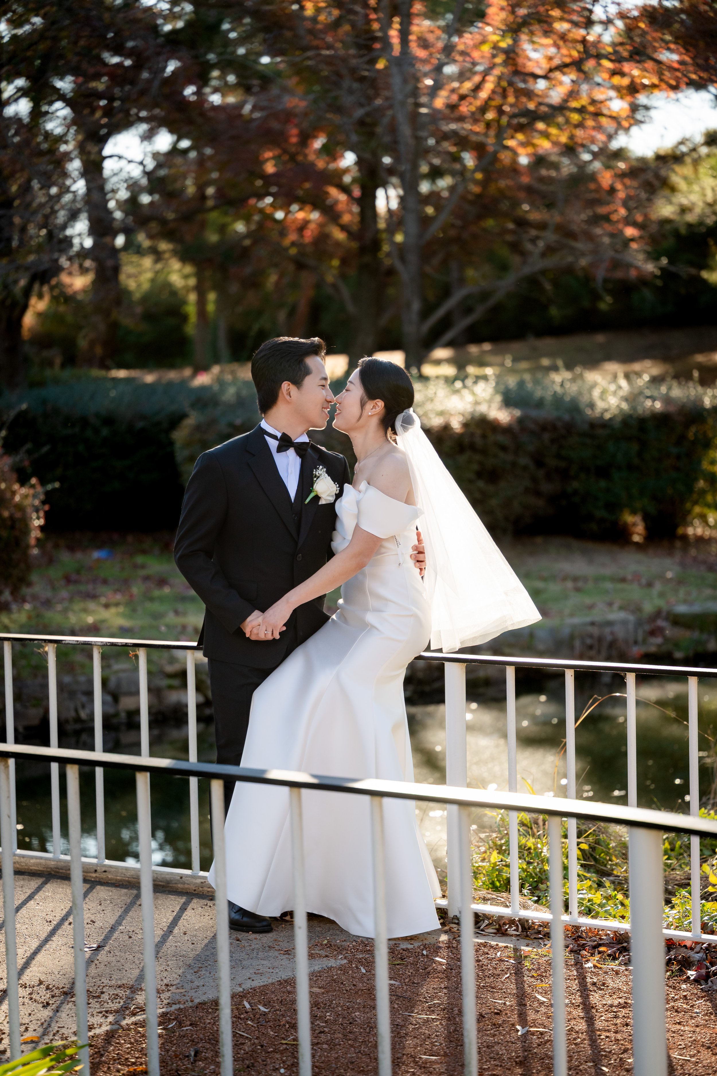 A bride and groom stand close together on a bridge in a park during fall, gazing into each other's eyes with trees and autumn foliage in the background.