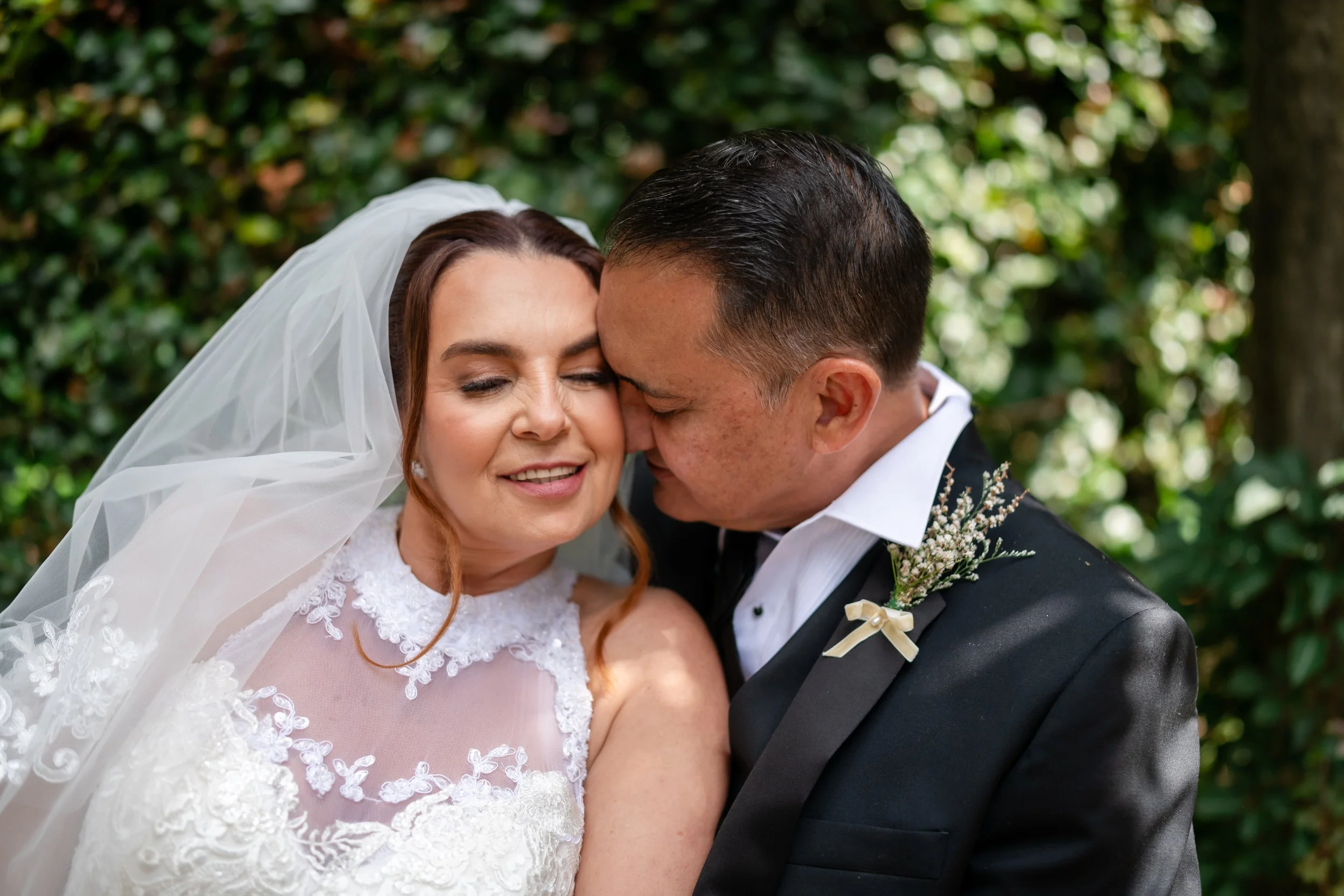A bride and groom sharing a kiss with foreheads touching, outdoors with greenery in the background.