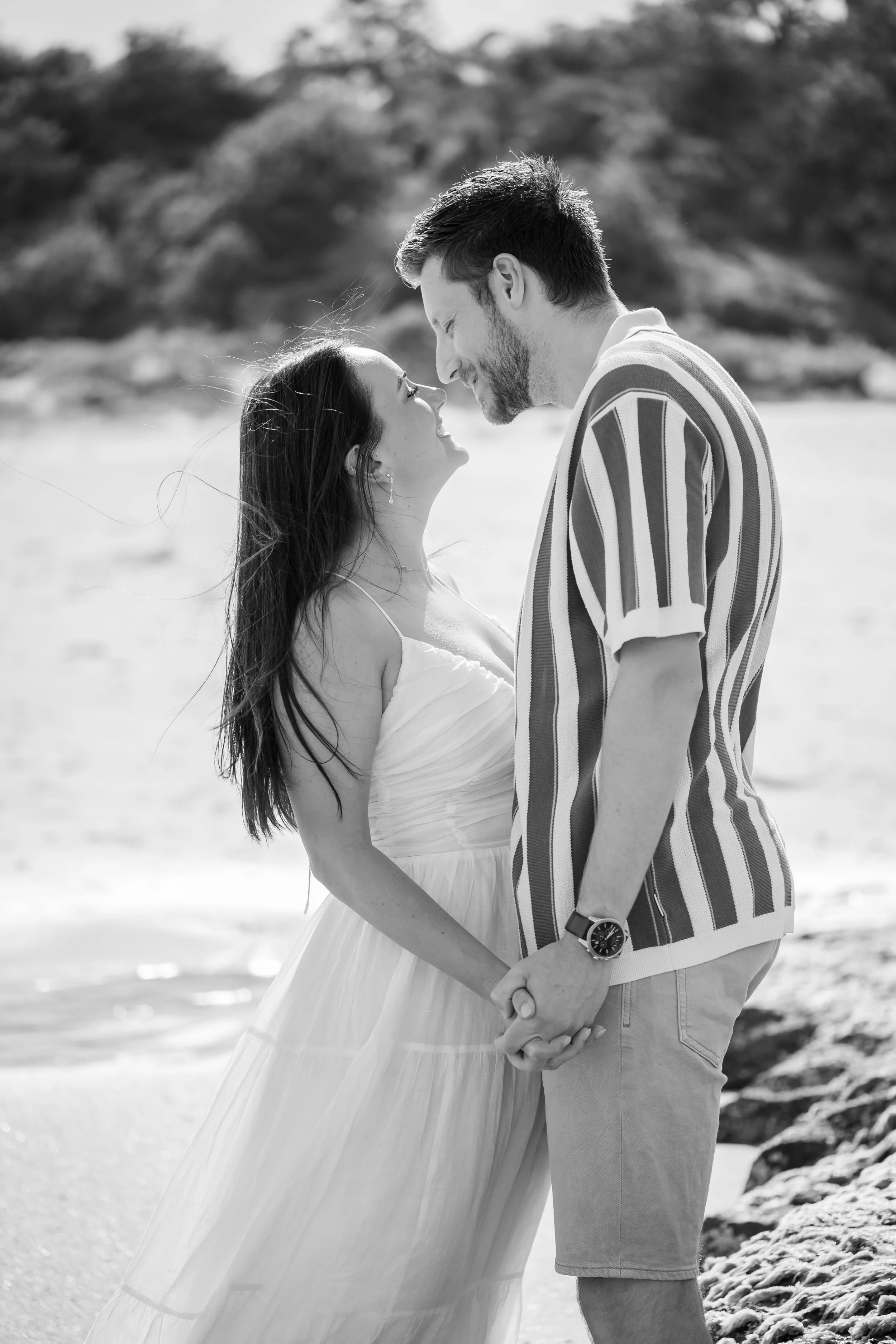 A black and white photo of a couple holding hands on a beach, gazing at each other, with plaques of rocks and ocean in the background.
