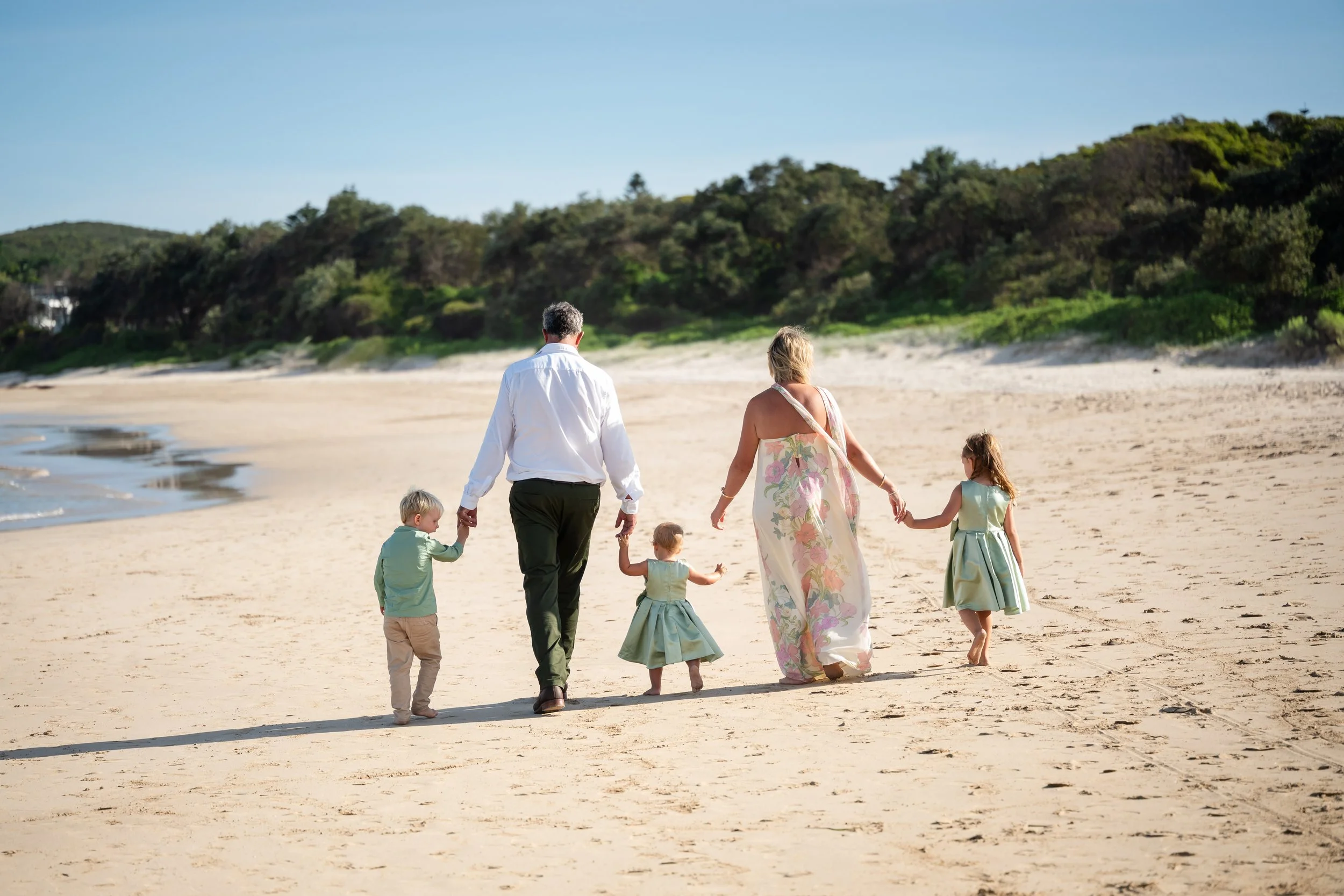 A family of five walking on a sandy beach holding hands, with trees in the background and clear blue sky.