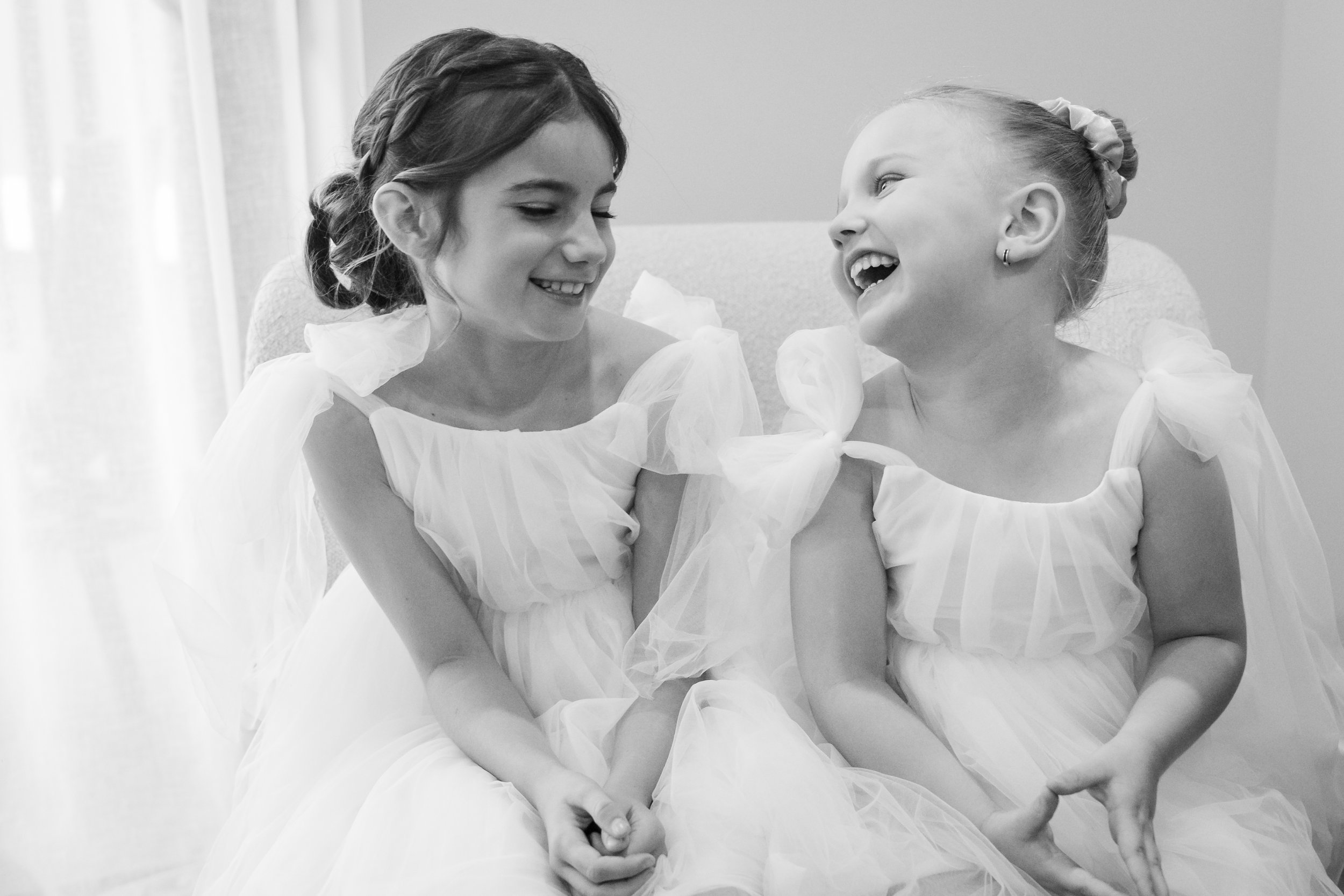 Two young girls in white dresses sitting on a couch, smiling and laughing at each other.