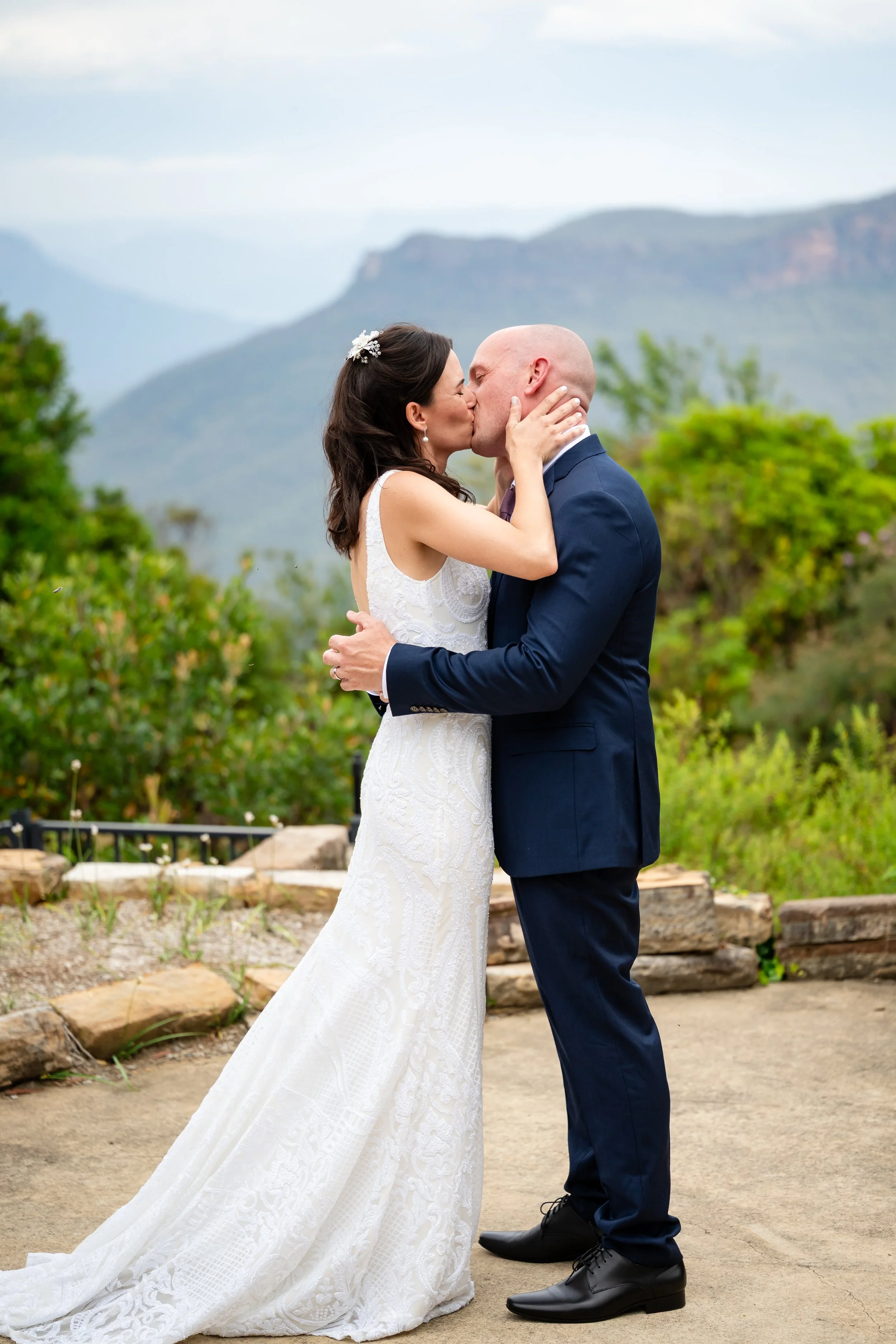A bride and groom kissing outdoors with a mountainous background.