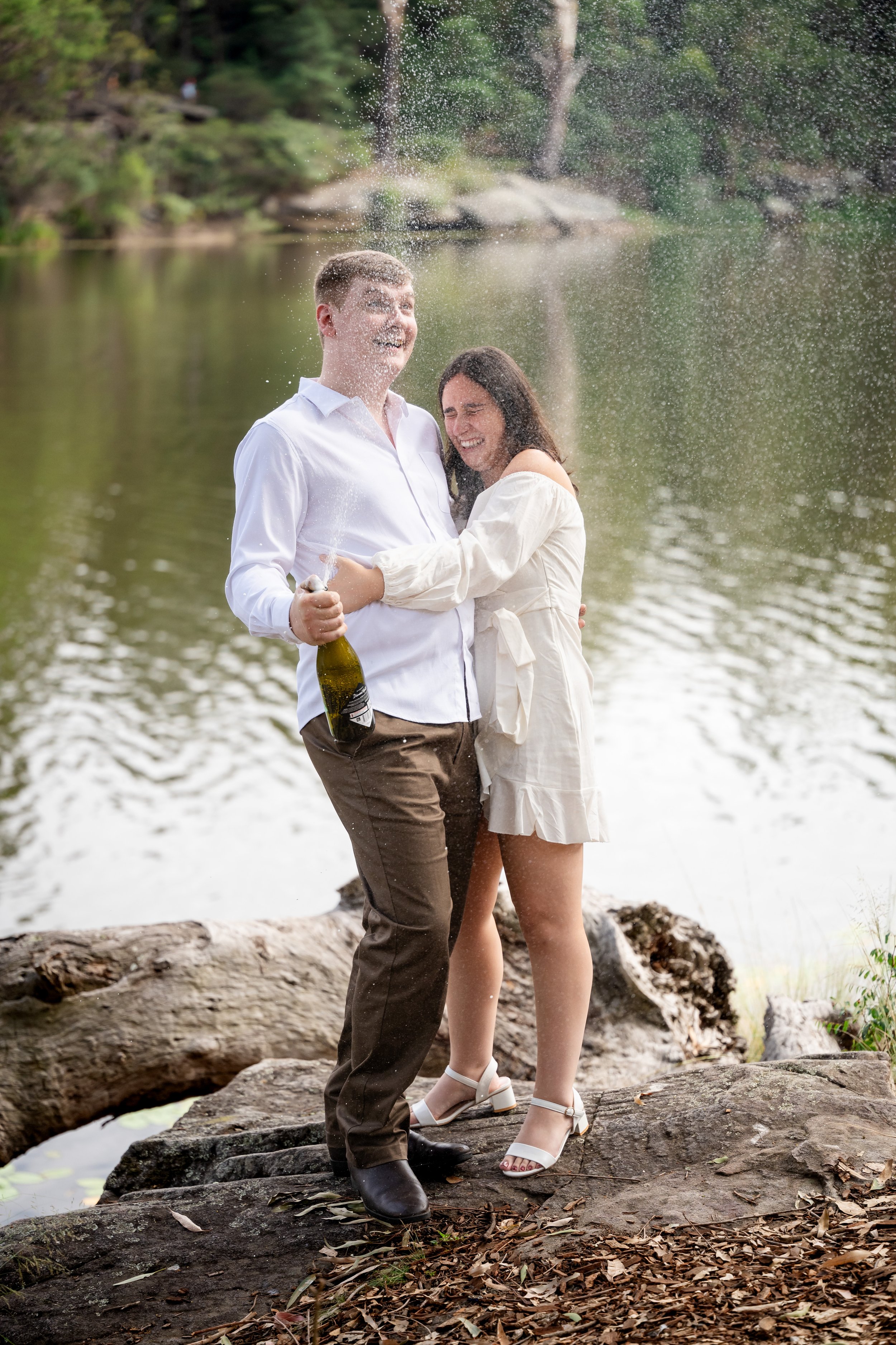 A couple celebrating near a lake, with the man holding a bottle of champagne and both smiling amid splashes of water.