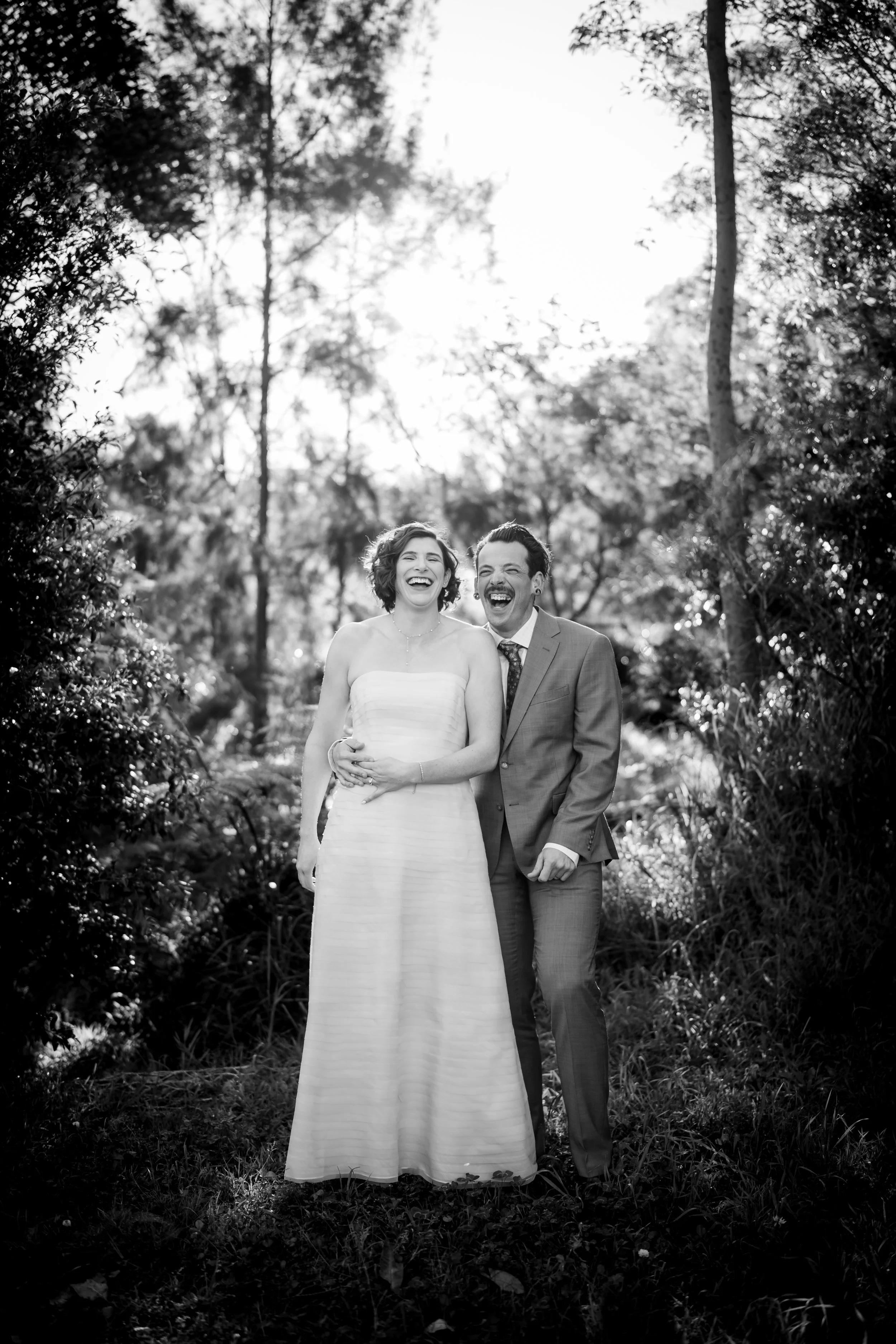 Black and white photo of a smiling bride in a strapless wedding dress and a smiling groom in a suit standing outdoors in a wooded area.
