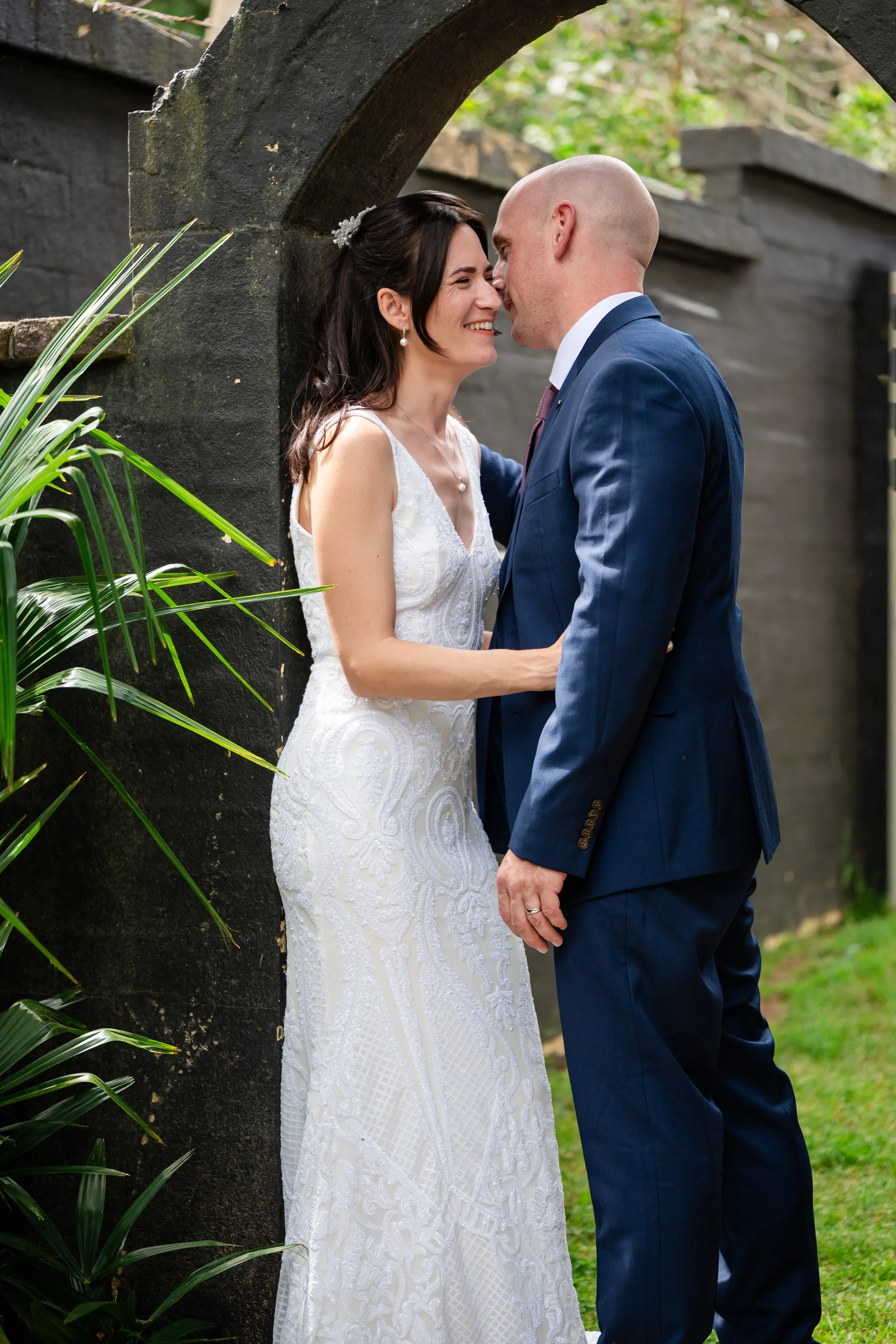 A bride and groom share an intimate moment during their wedding, standing close together outdoors near a stone archway, smiling and touching noses.