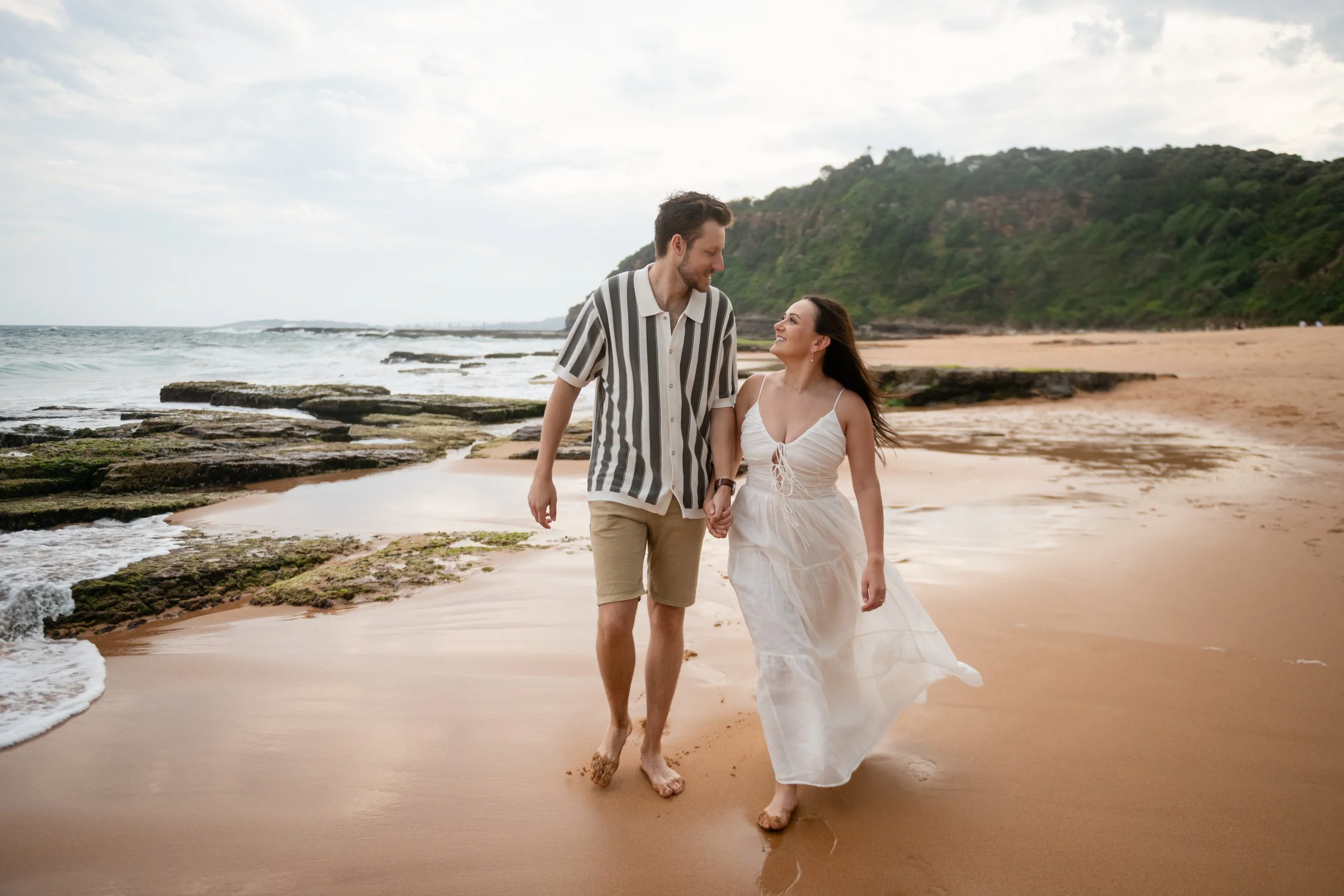 A couple holding hands and walking on a beach with rocks and green hills in the background