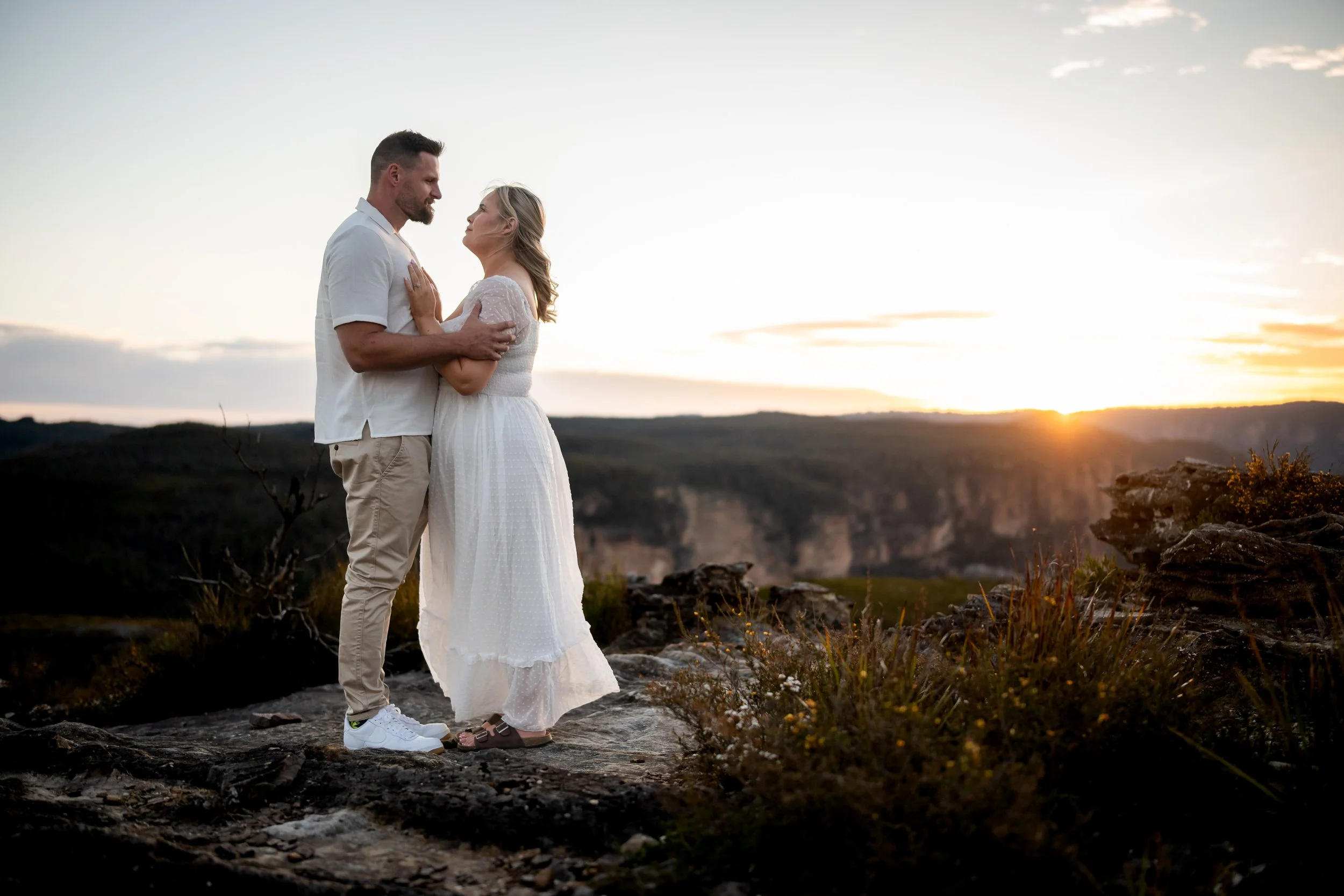 A couple standing on rocks at sunset, embracing each other, with a scenic canyon in the background.