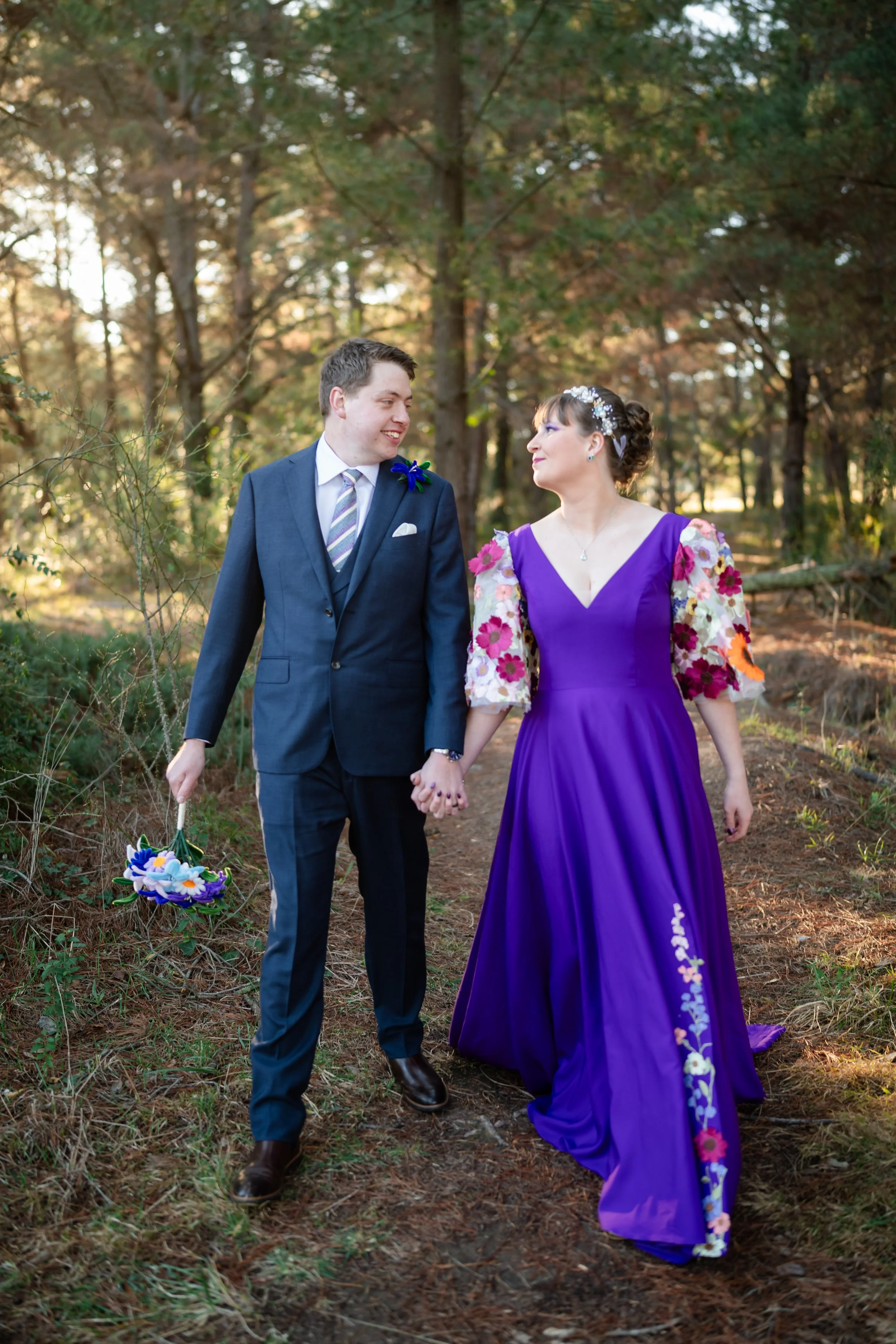 A couple holding hands walking on a forest trail, smiling at each other. The man is wearing a navy suit with a striped tie and holding a small bouquet, while the woman is in a purple dress with floral embroidered sleeves and a floral headpiece.
