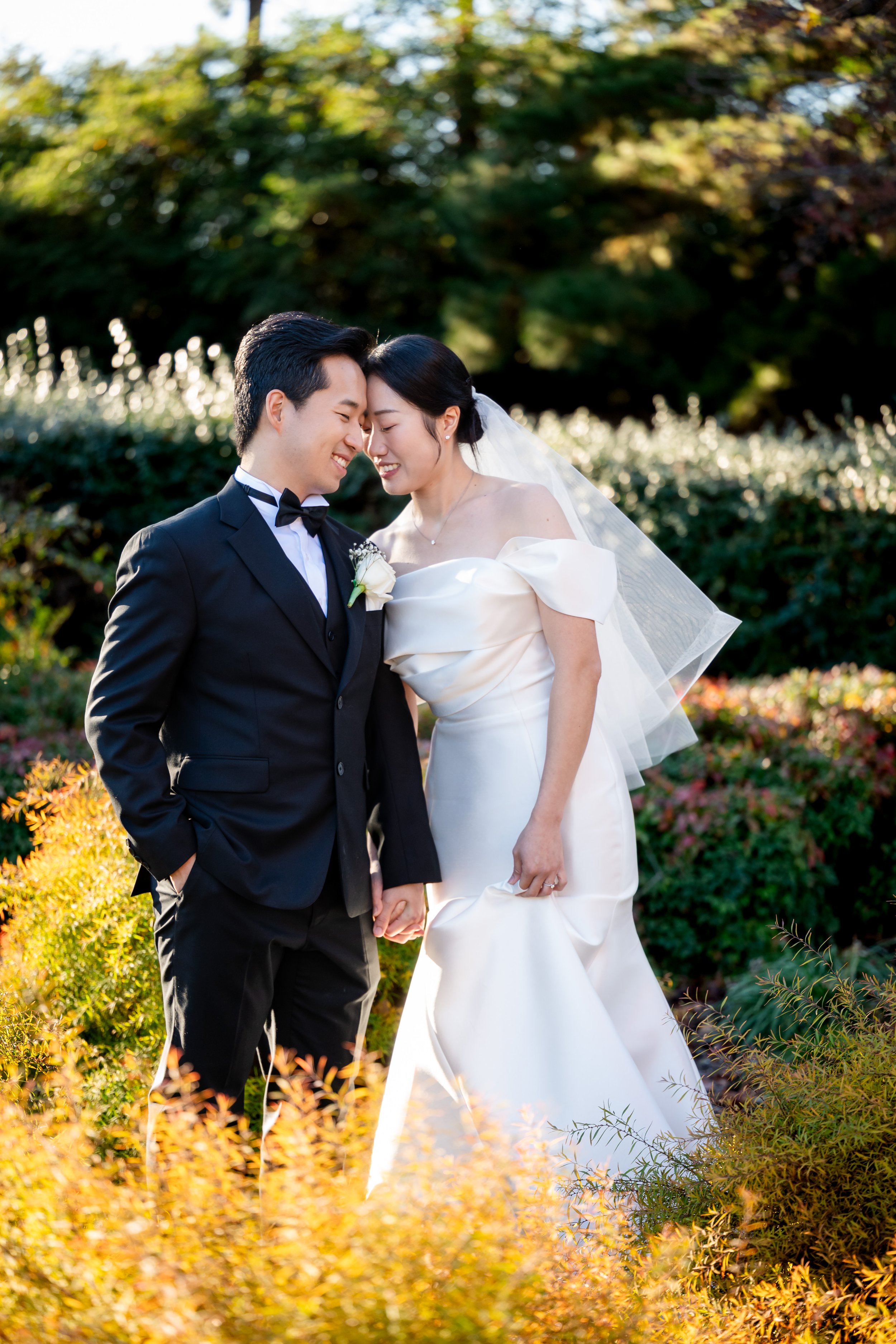 A newlywed couple in wedding attire hugging and smiling with foreheads touching outdoors, surrounded by vibrant autumn foliage.