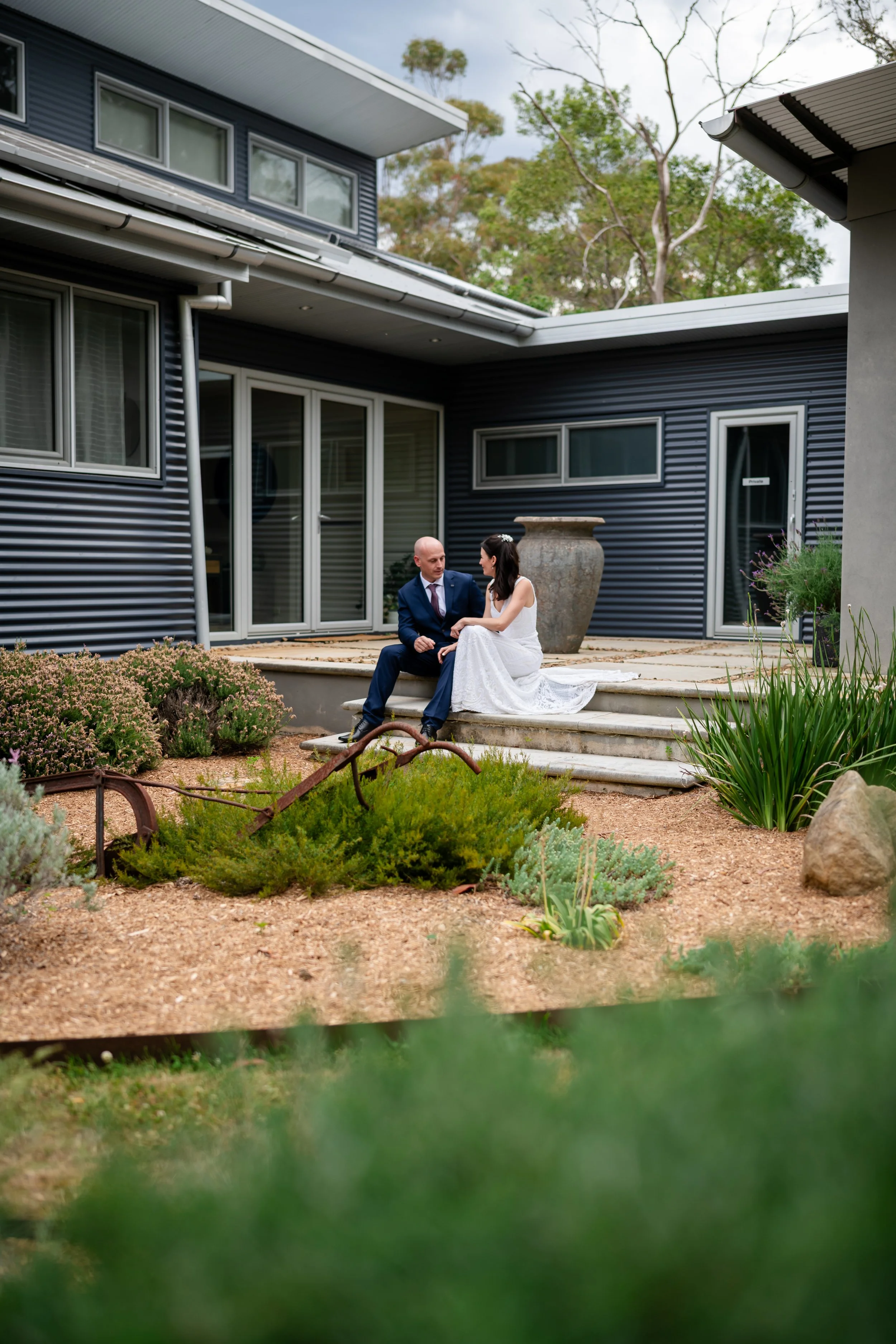 A newlywed couple sitting on steps outside a modern house, with wedding attire, surrounded by a landscaped garden.
