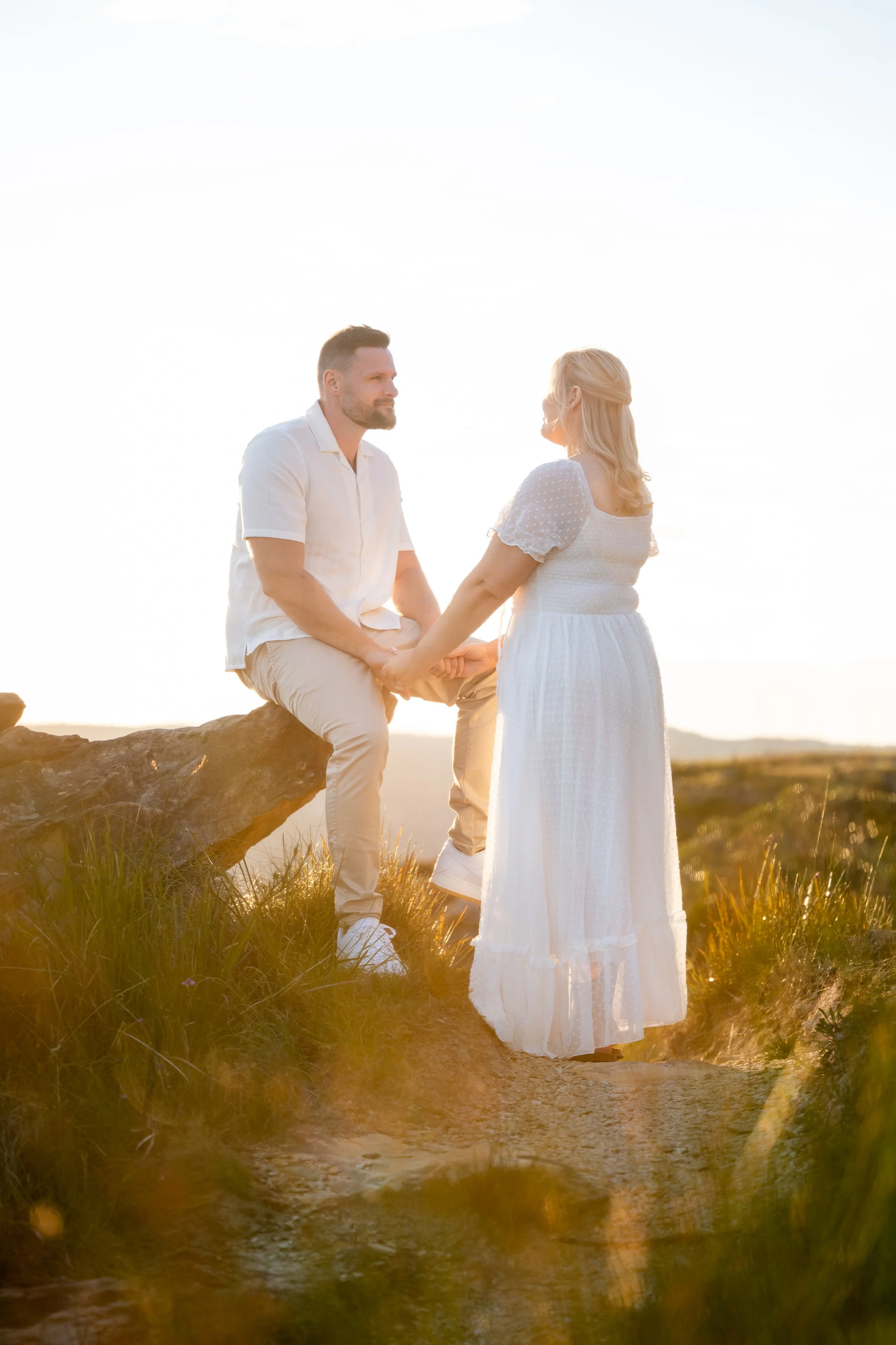 A couple holding hands outdoors at sunset, with the man sitting on a rock and the woman standing on a dirt path, both dressed in white.