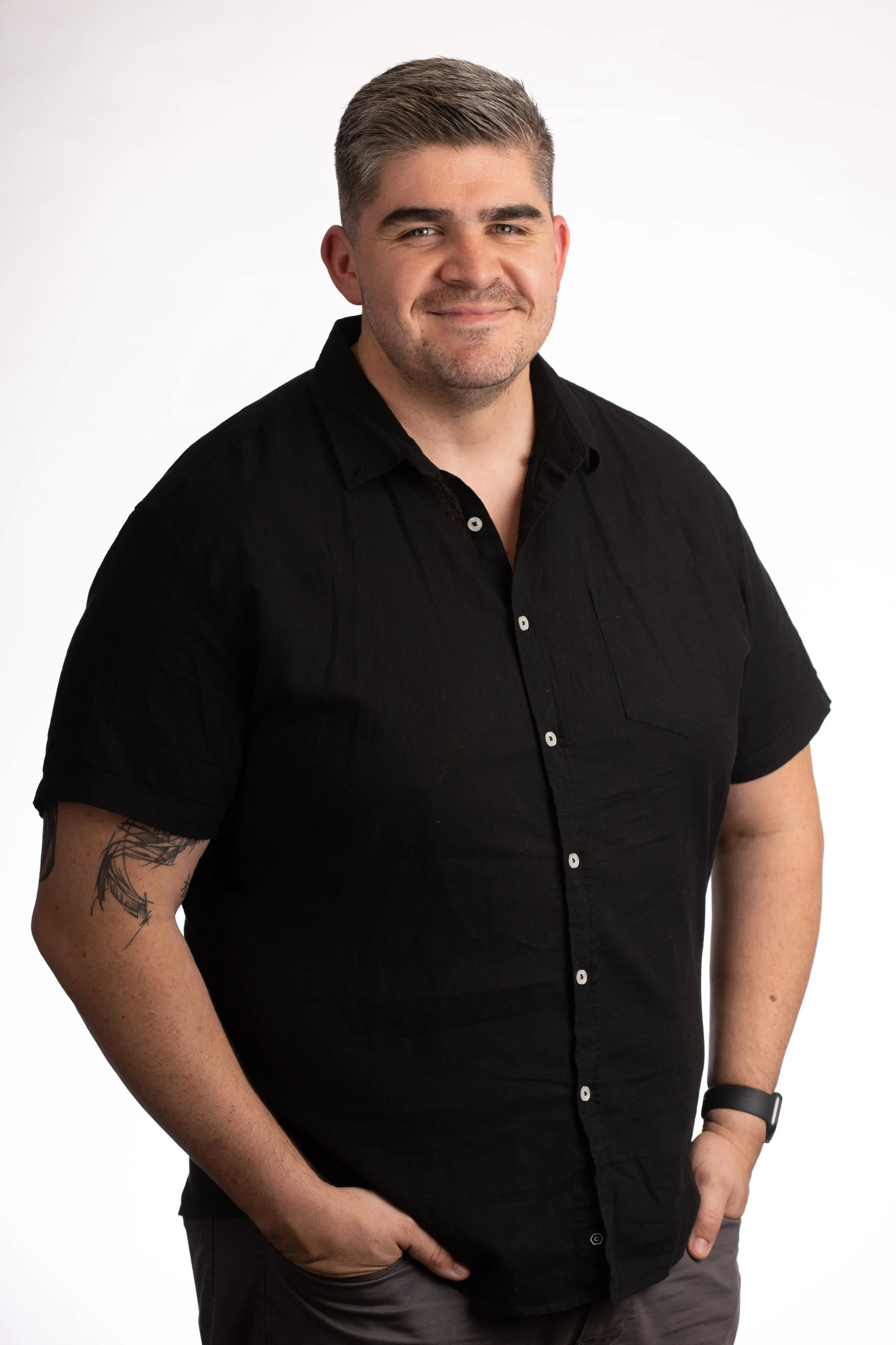 A man with short gray hair, wearing a black button-up shirt and a black watch on his left wrist, standing against a plain white background.