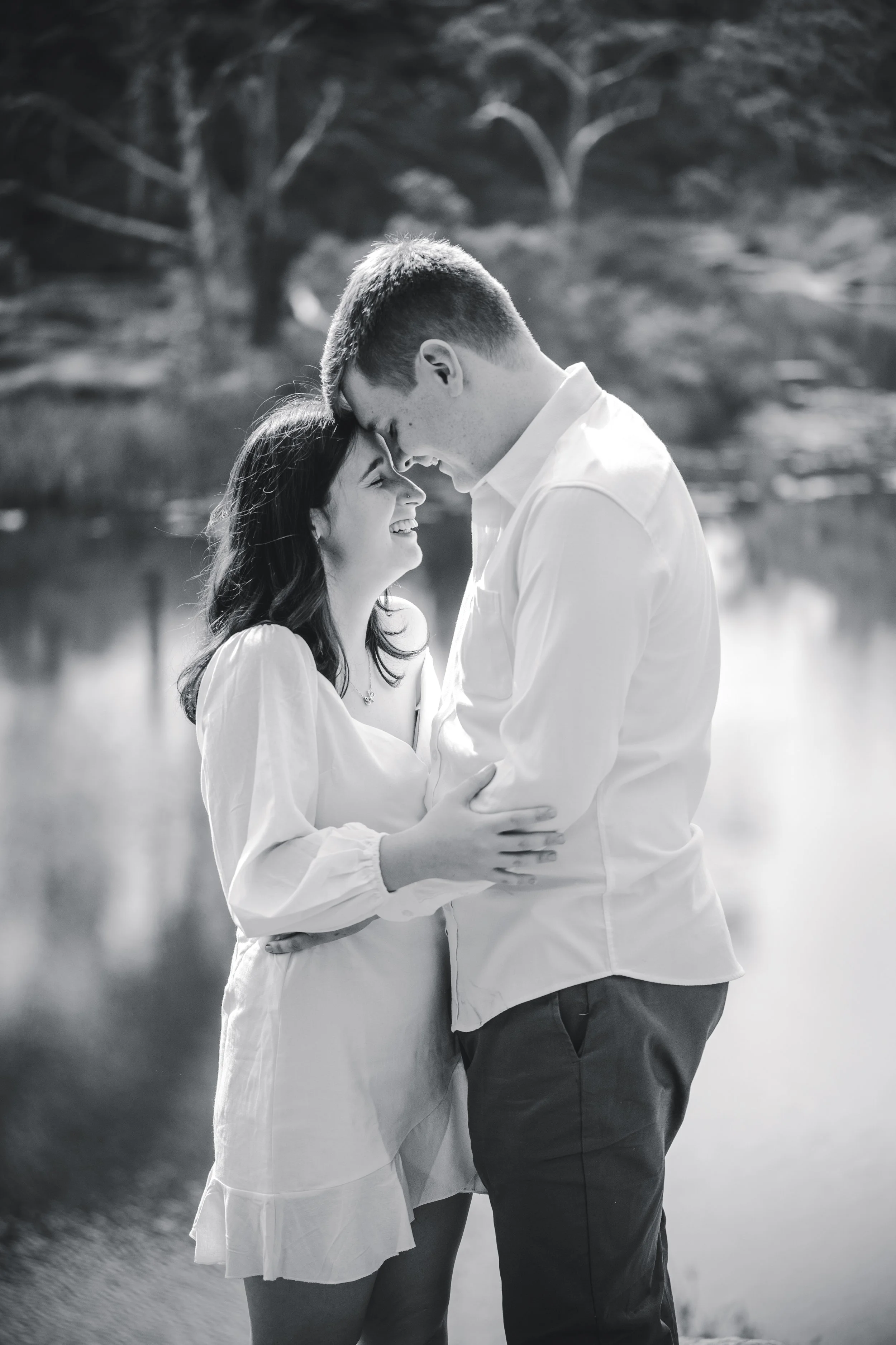 A black and white photo of a couple standing near a lake, smiling with foreheads touching, holding each other affectionately.
