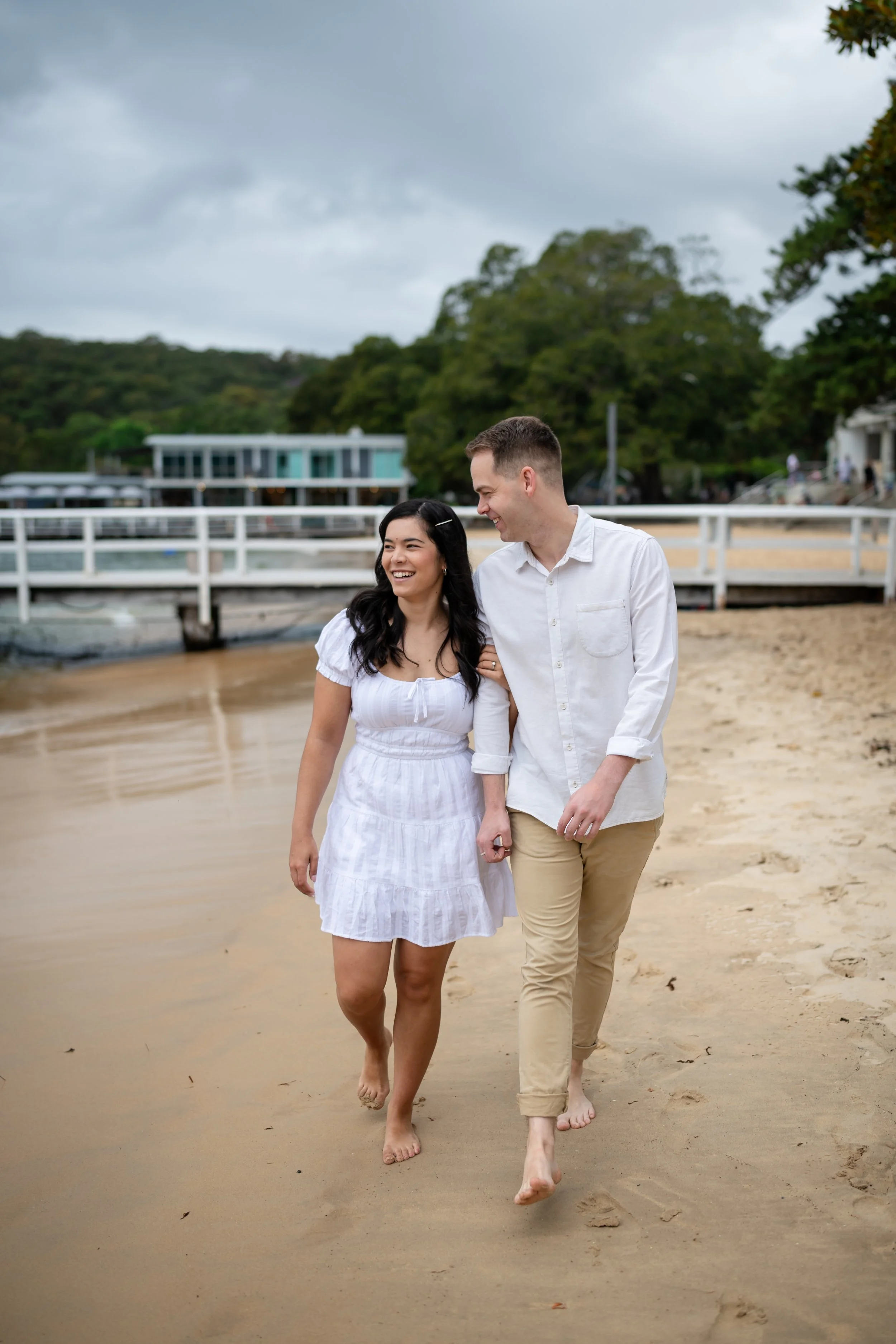 A smiling young couple walking barefoot on a sandy beach, holding hands, with a pier, a building, and lush green trees in the background under a cloudy sky.
