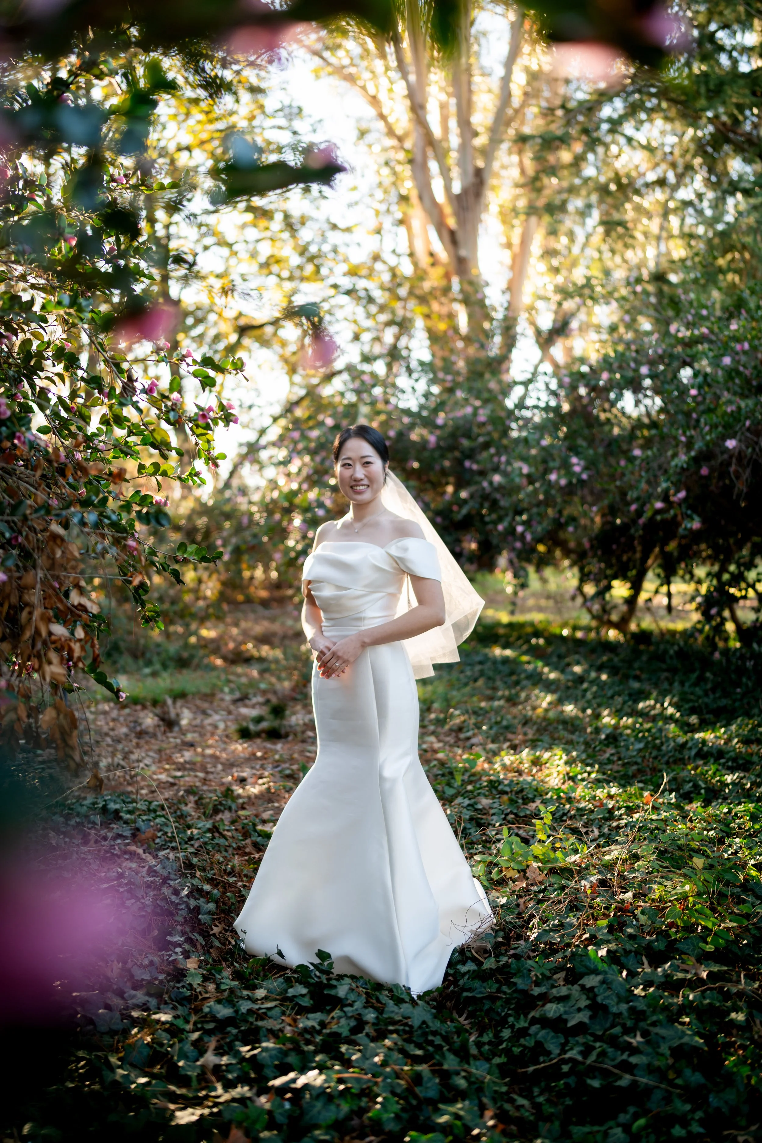A bride standing outdoors in a garden, wearing a white wedding dress with off-the-shoulder sleeves and a veil, smiling at the camera, with sunlight filtering through trees and bushes around her.