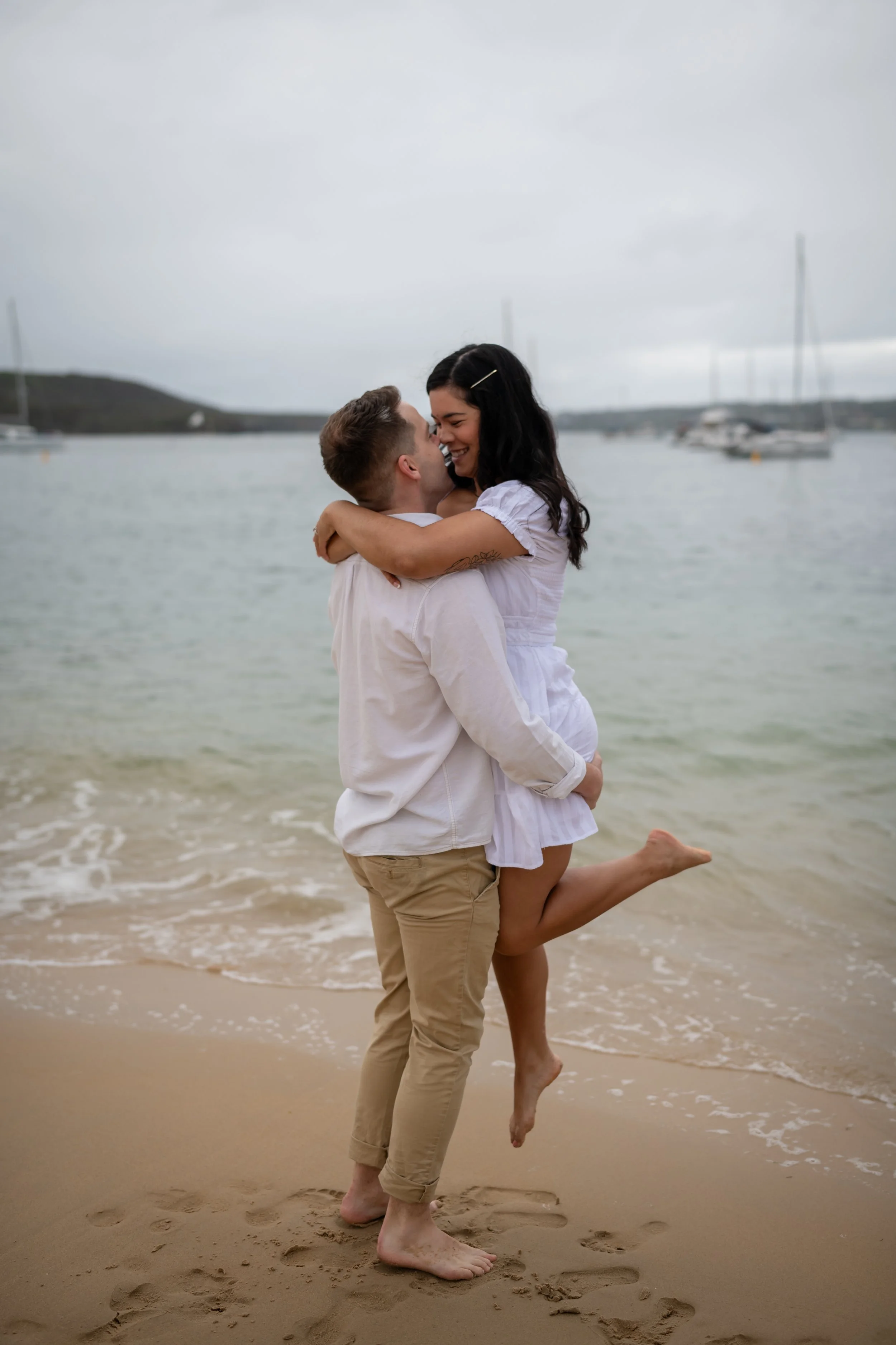 A couple embracing on the beach, with the woman lifting her leg, near the water with boats in the background.