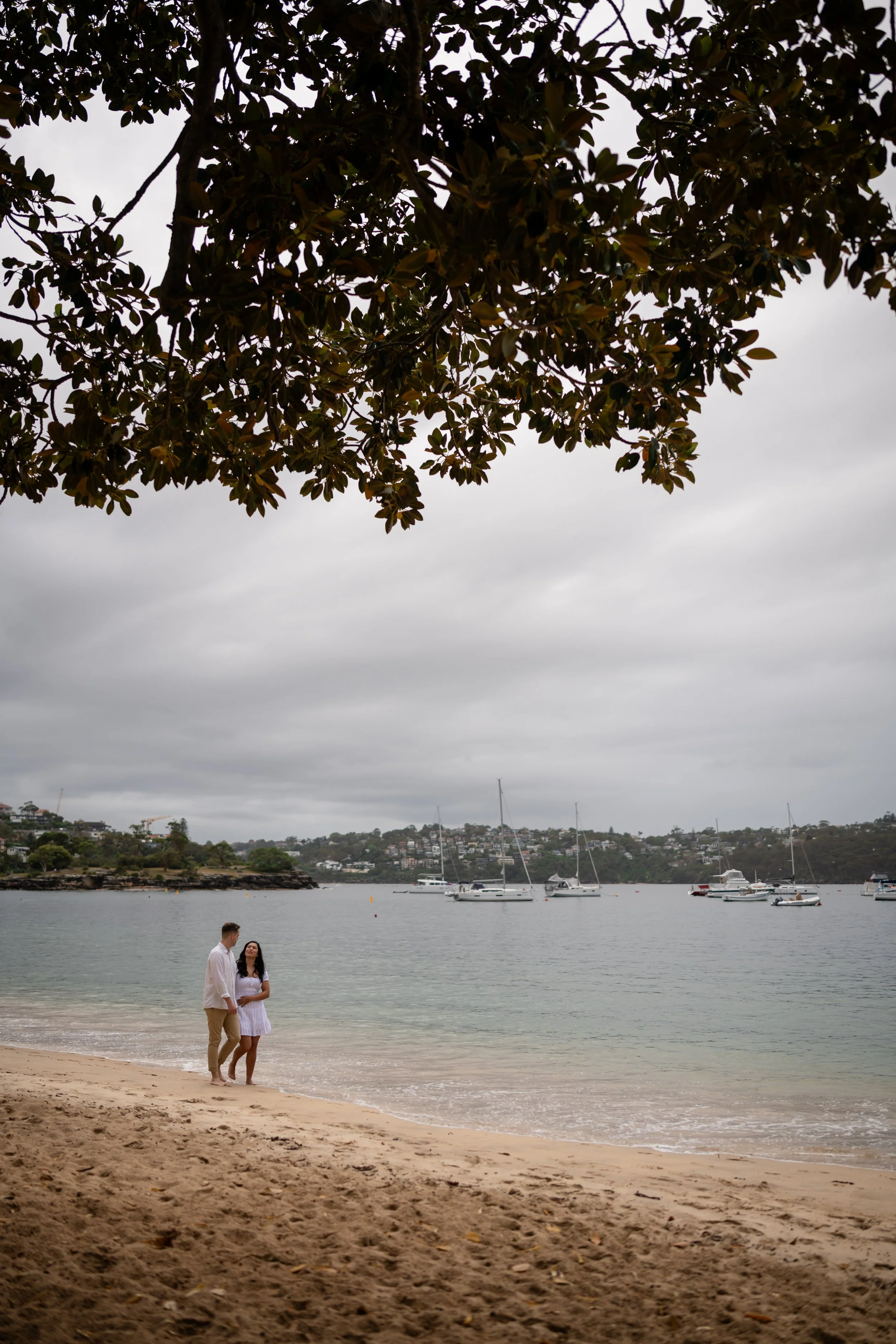 A couple standing on a sandy beach near the water under cloudy skies, with boats anchored in the bay and a hillside with houses in the background.