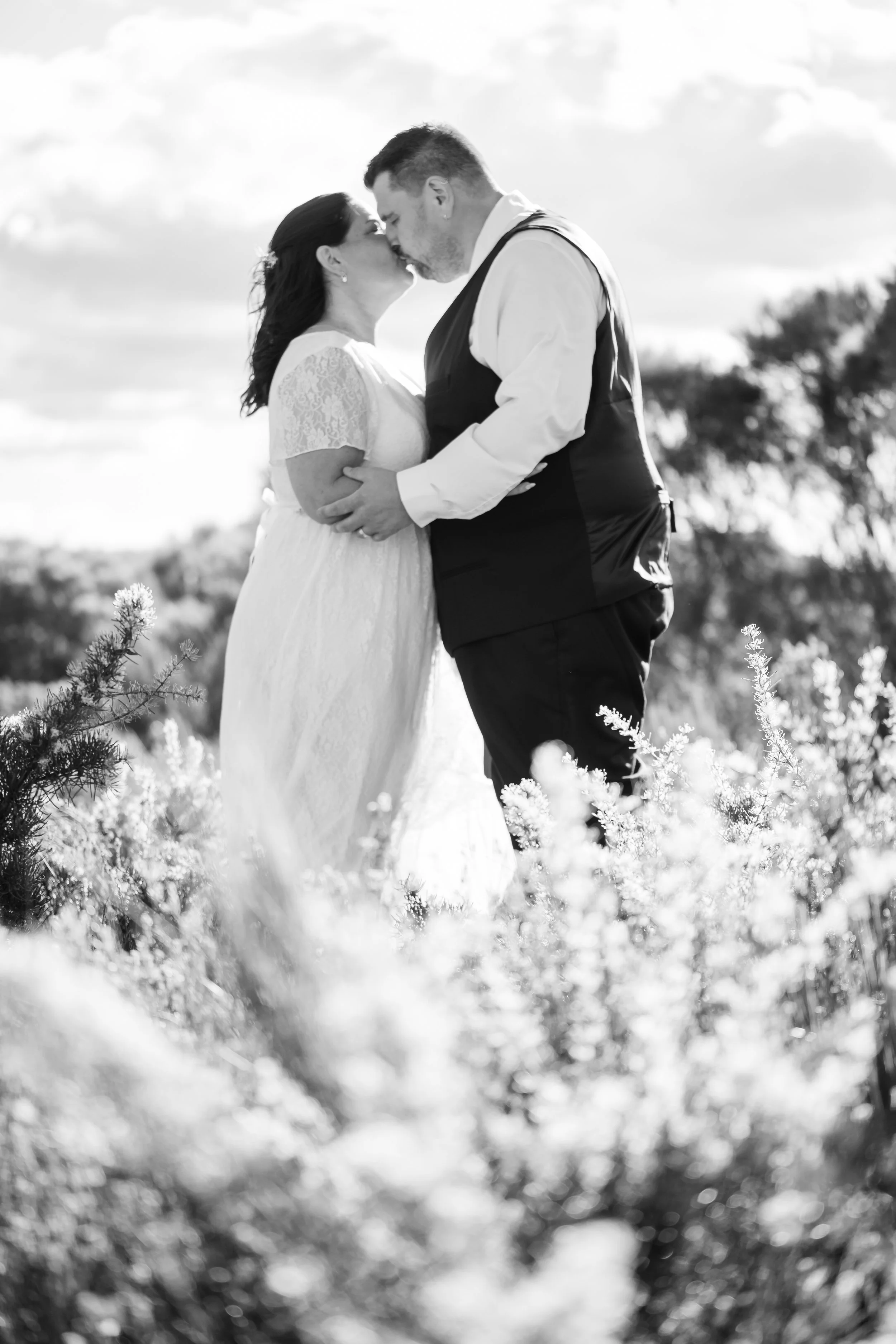 Black and white photo of a bride and groom kissing outdoors with flowers and sky in the background.