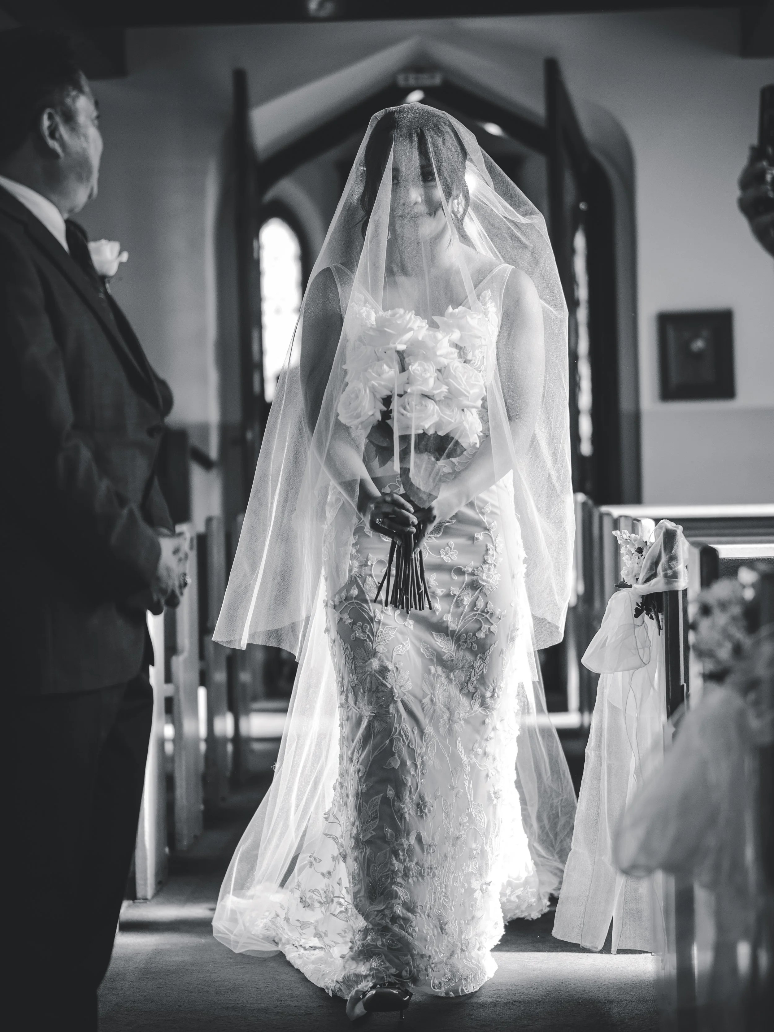 A bride in a wedding dress with floral embroidery holding a bouquet, walking down the aisle inside a church with a veil covering her face, smiling, during a wedding ceremony.