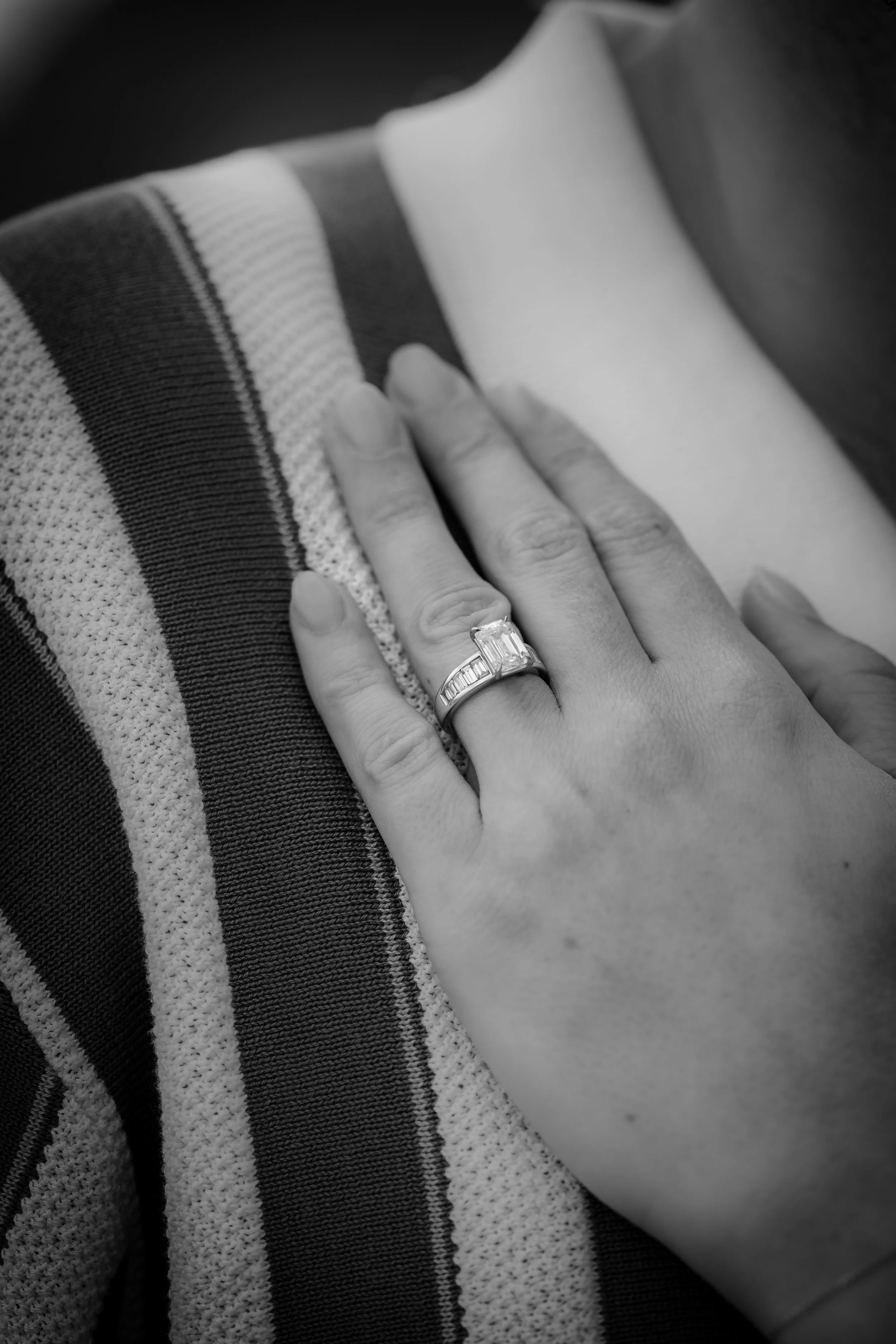 Close-up of a woman's hand wearing an engagement ring with a large rectangular diamond and a wedding band with smaller diamonds, resting on a striped fabric surface.
