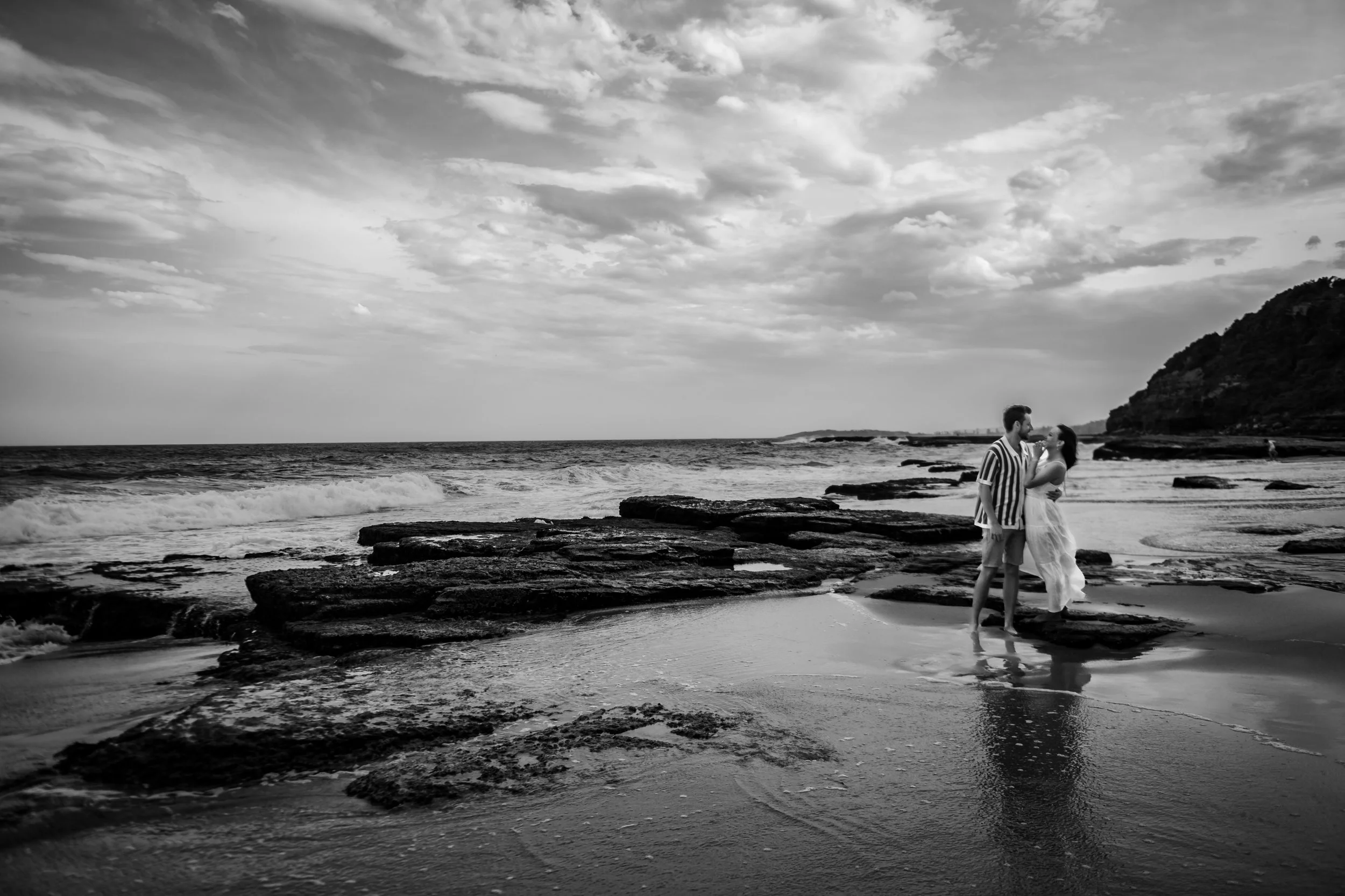 A couple standing on a rocky beach, sharing a kiss near the water, with a cliff in the background under cloudy skies in black and white.