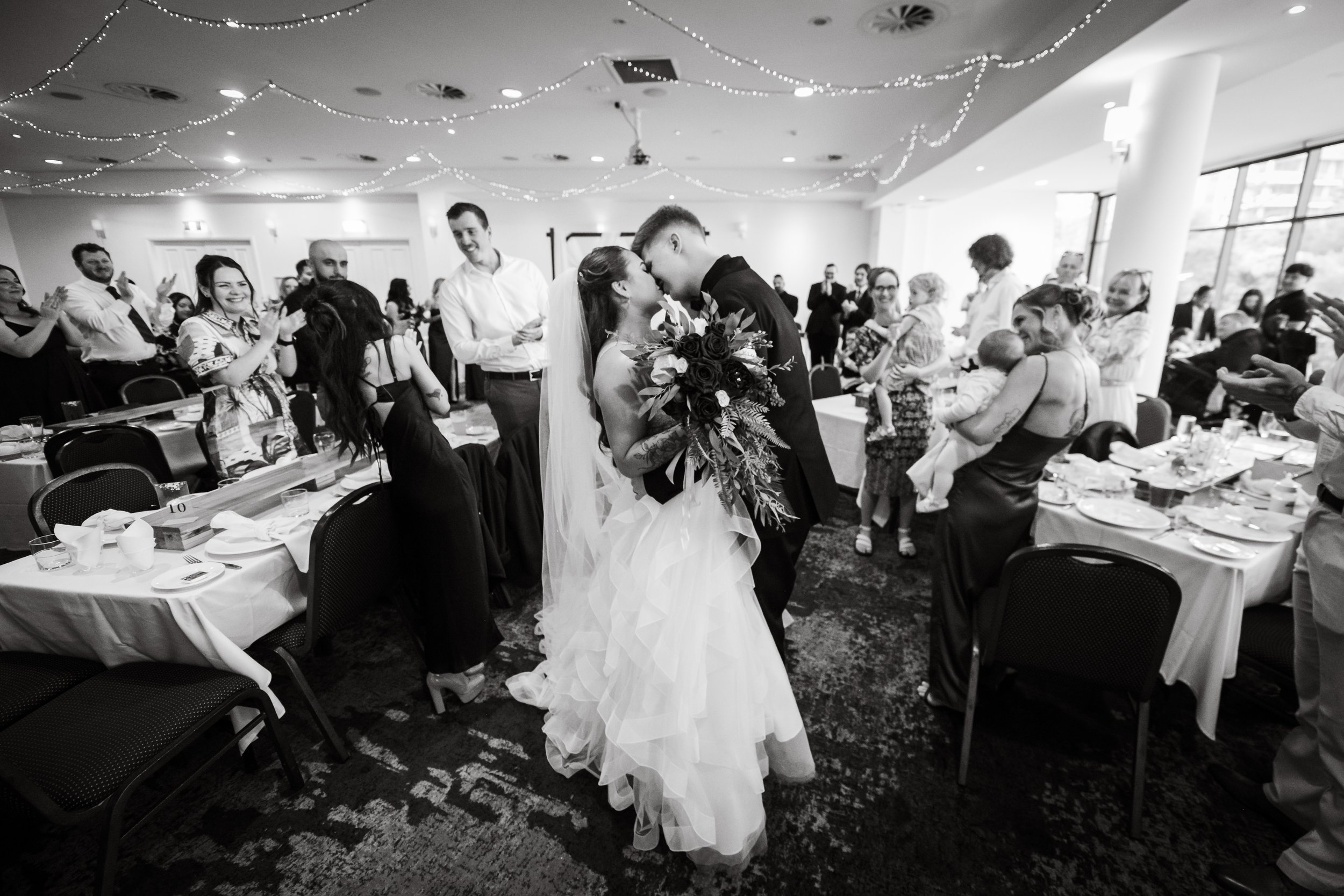 A newlywed couple is sharing a kiss at their wedding reception, surrounded by guests clapping and smiling.