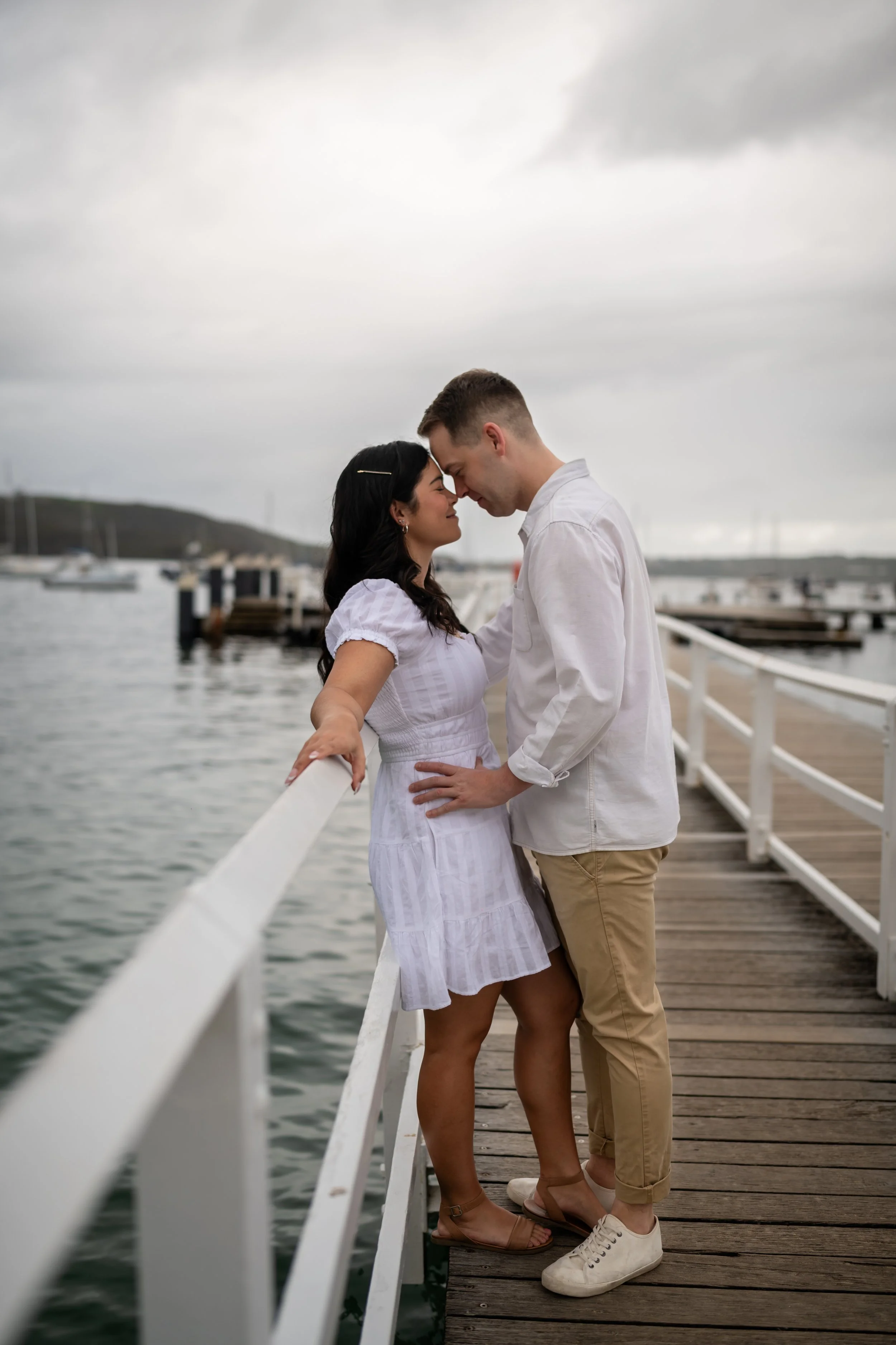 A couple stands close on a wooden dock by water, touching foreheads, with boats in the background and overcast sky.