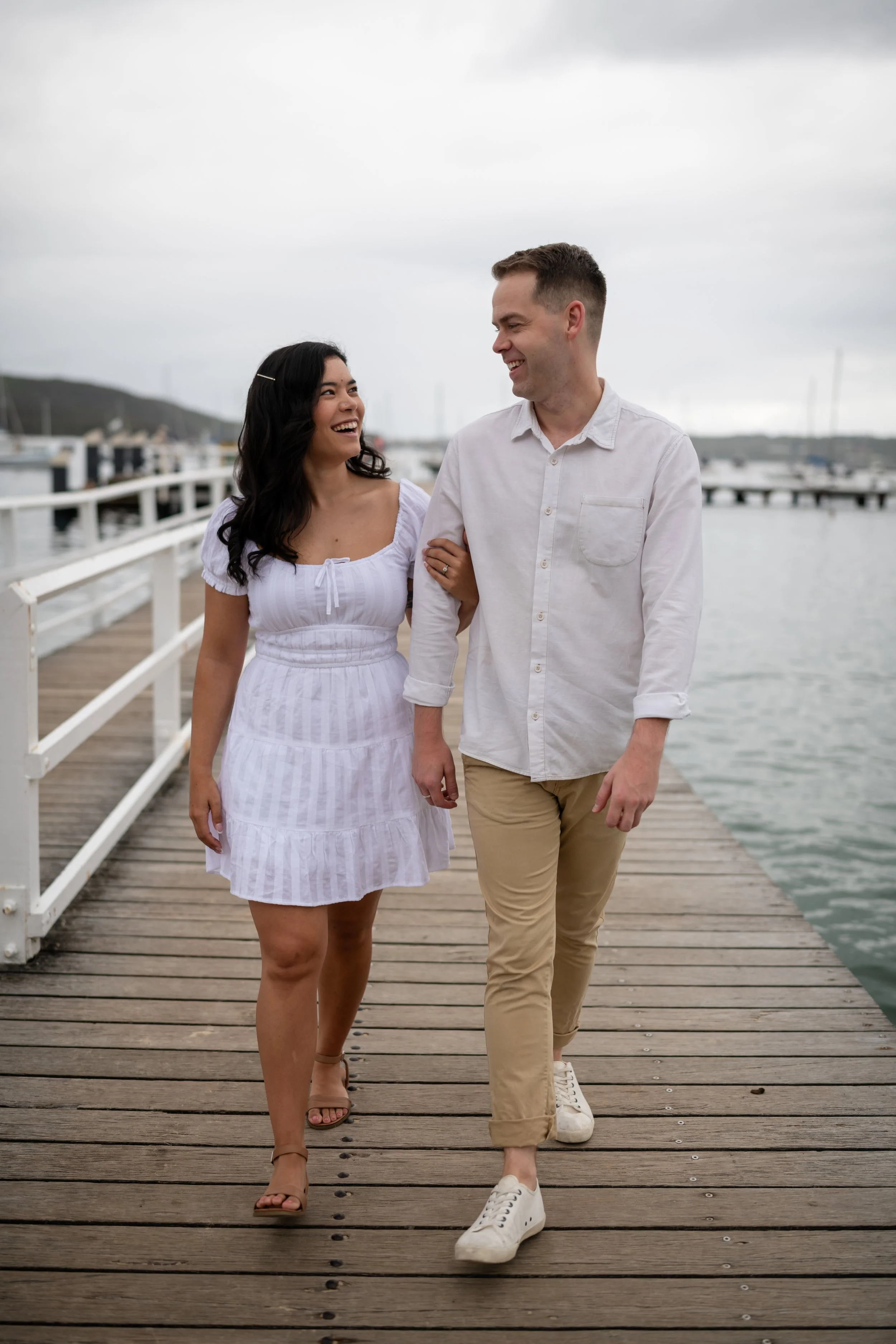 A couple walks arm in arm on a wooden dock by the water, smiling at each other, with boats and a cloudy sky in the background.
