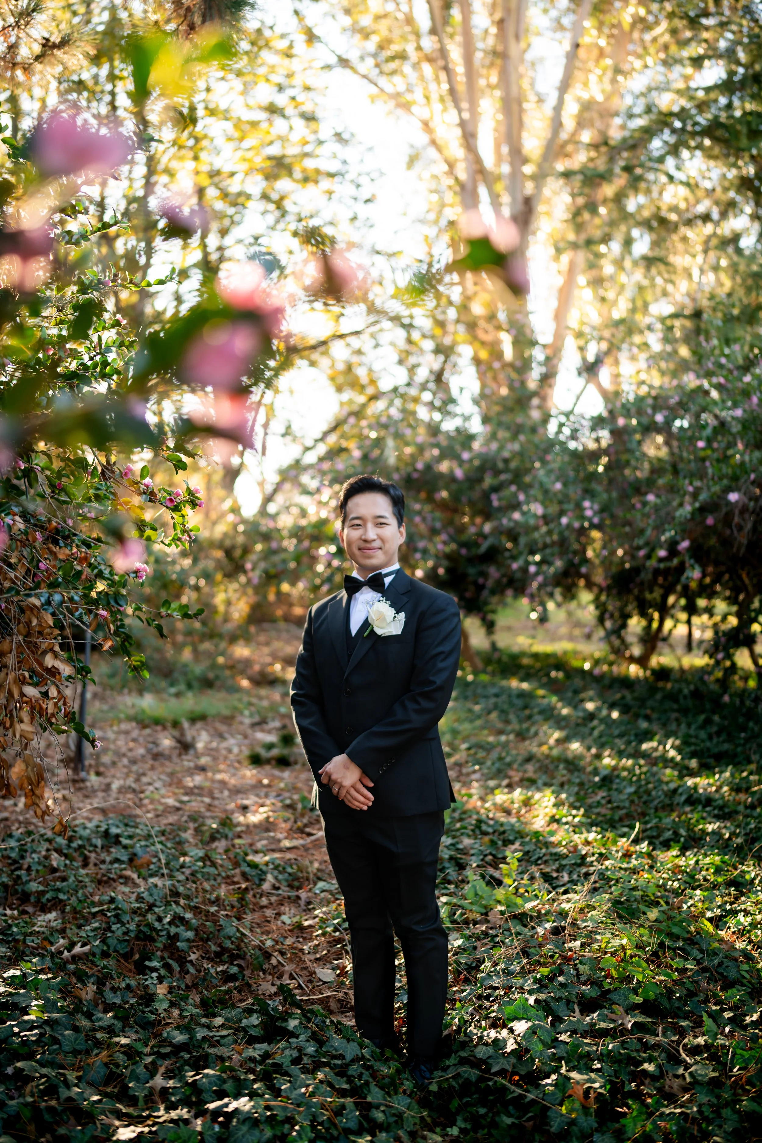 A man in a black tuxedo with a bowtie and white boutonniere stands outdoors among trees and flowering bushes, smiling at the camera during sunset.