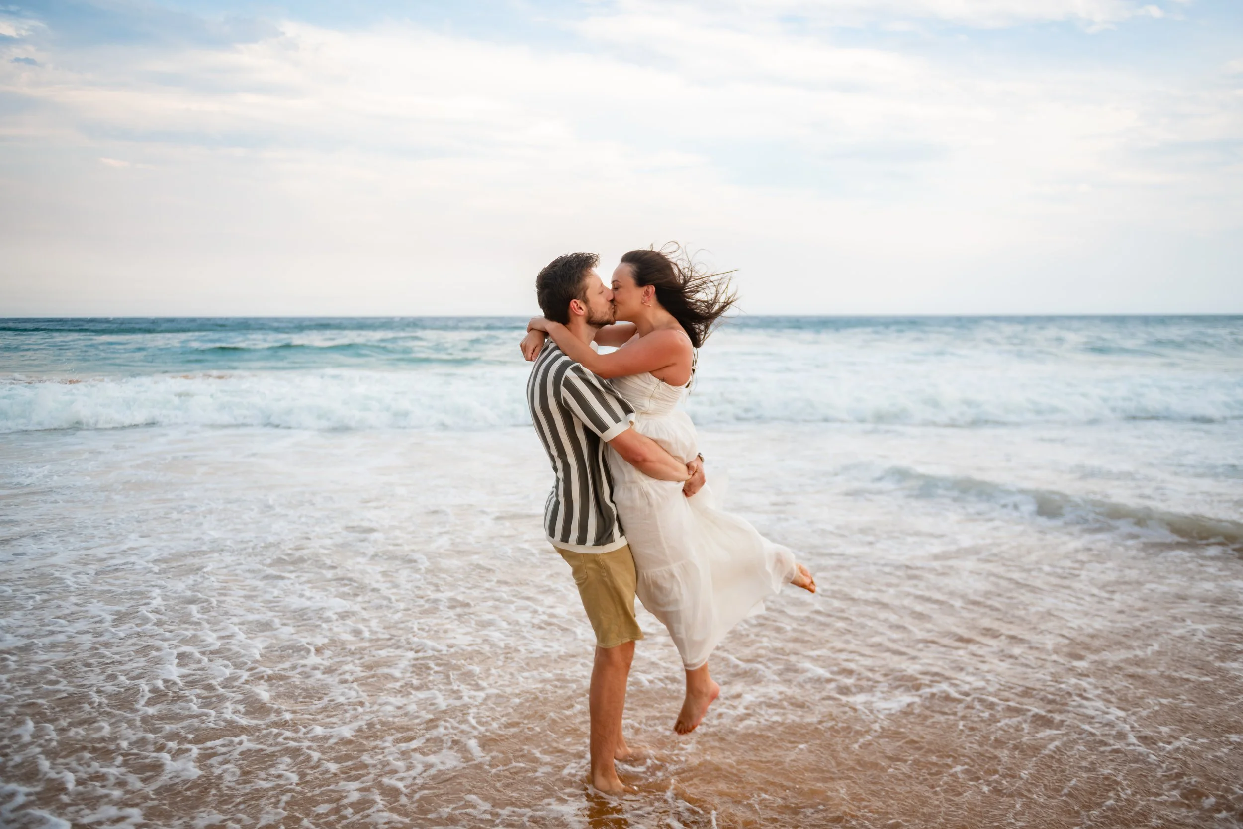 A man lifts a woman in a white dress on the beach as they kiss, with ocean waves and a cloudy sky in the background.