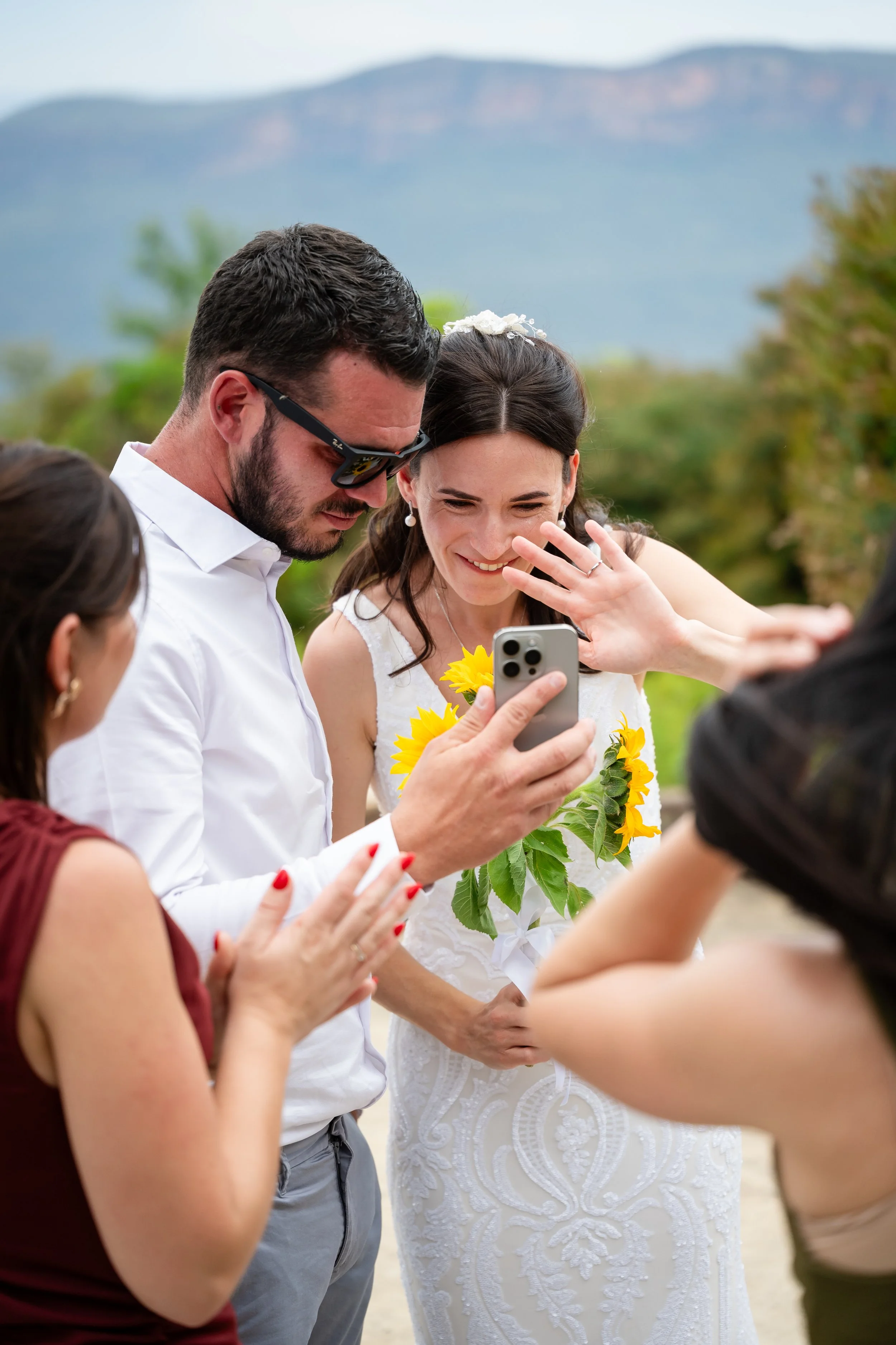A bride in a white wedding dress holding sunflowers, smiling as friends take a photo of her outdoors with a scenic mountain background.