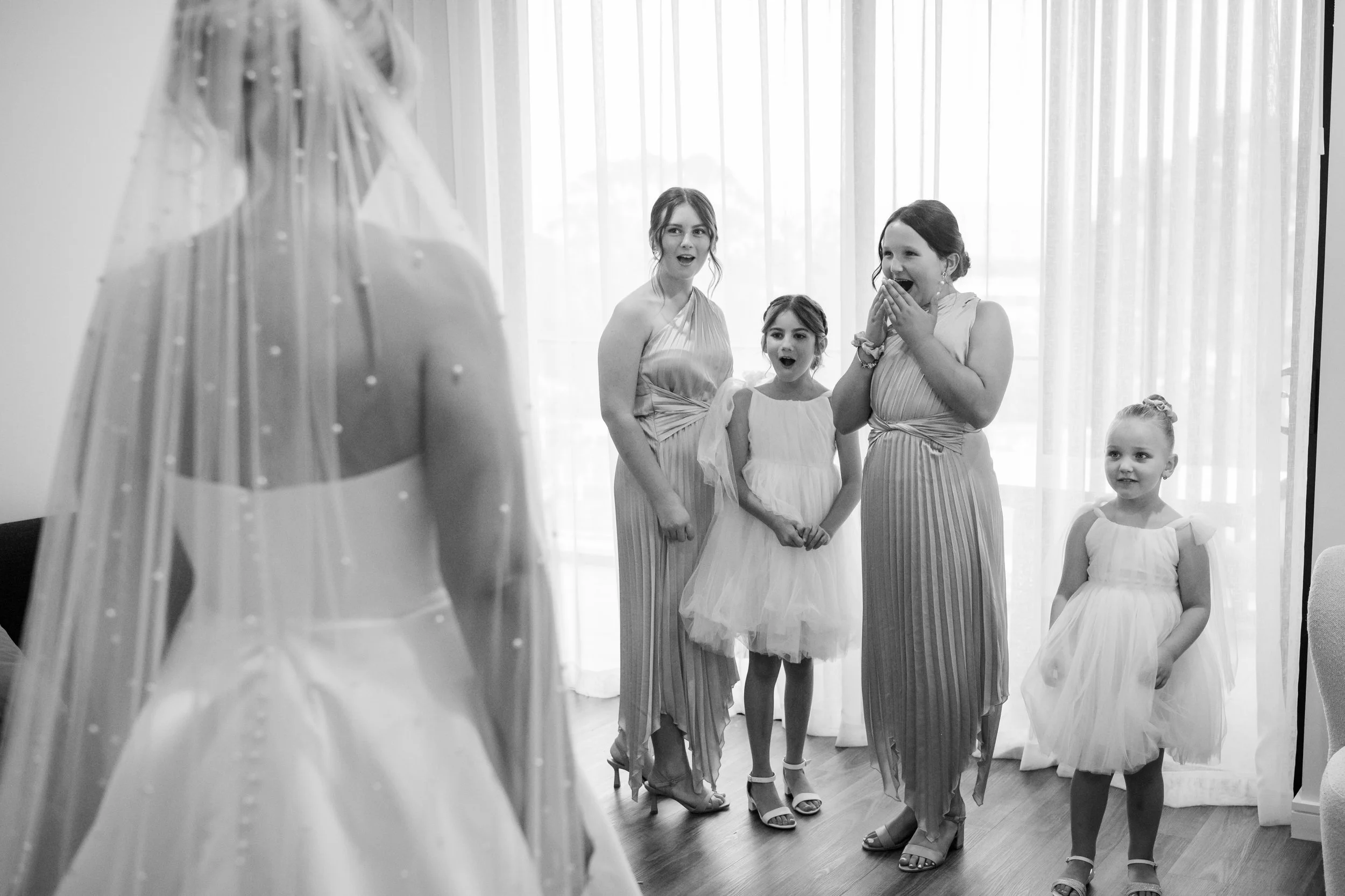 A bride in a wedding dress with a veil facing a group of women and girls in fancy dresses, who look surprised and happy, indoors with white curtains in the background.