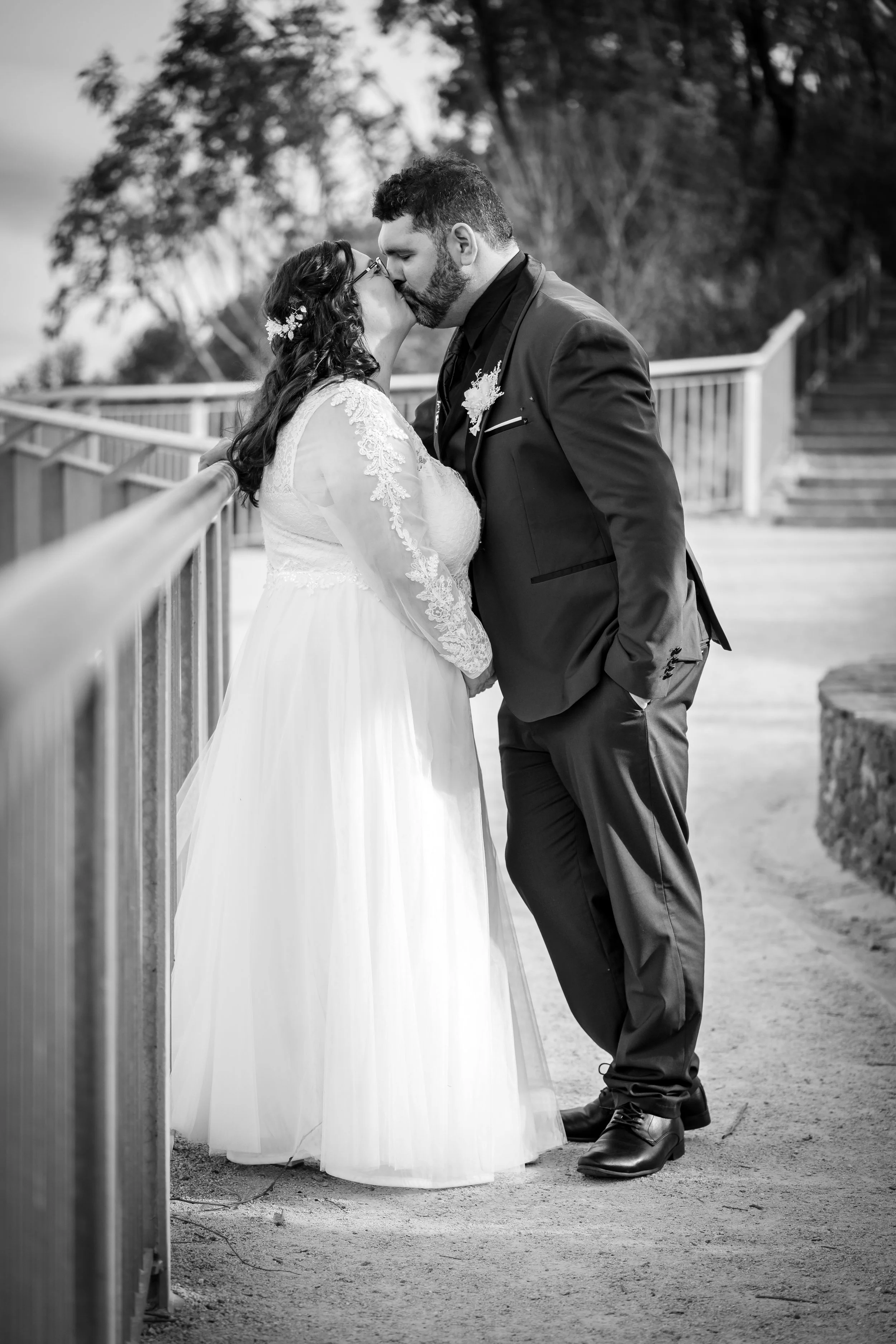 A black and white photo of a bride and groom sharing a kiss outdoors, with the bride in a lace wedding dress and the groom in a suit, standing next to a railing.
