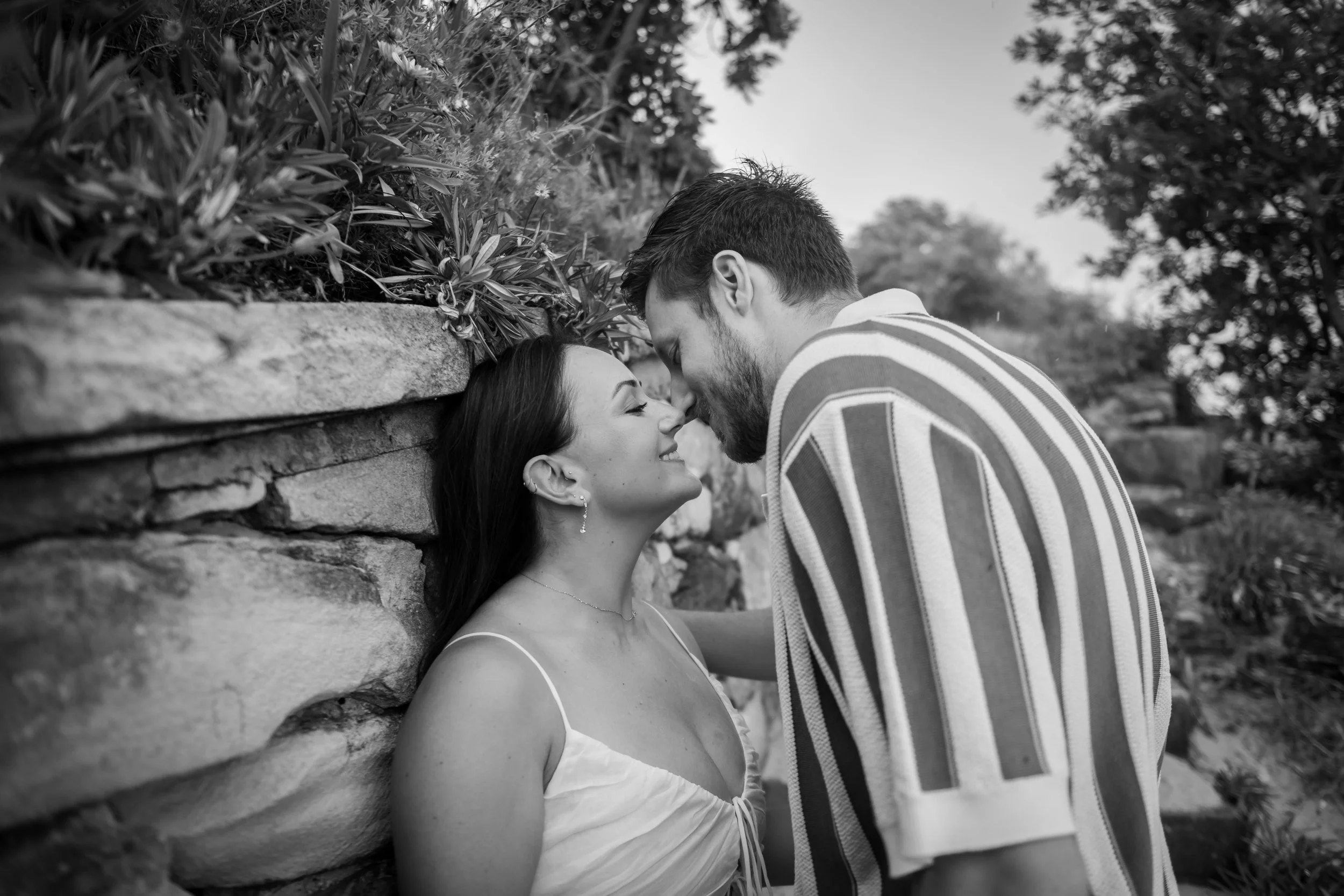 A black and white photo of a couple leaning close against a stone wall, about to kiss, with their foreheads touching, outdoors with trees in the background.