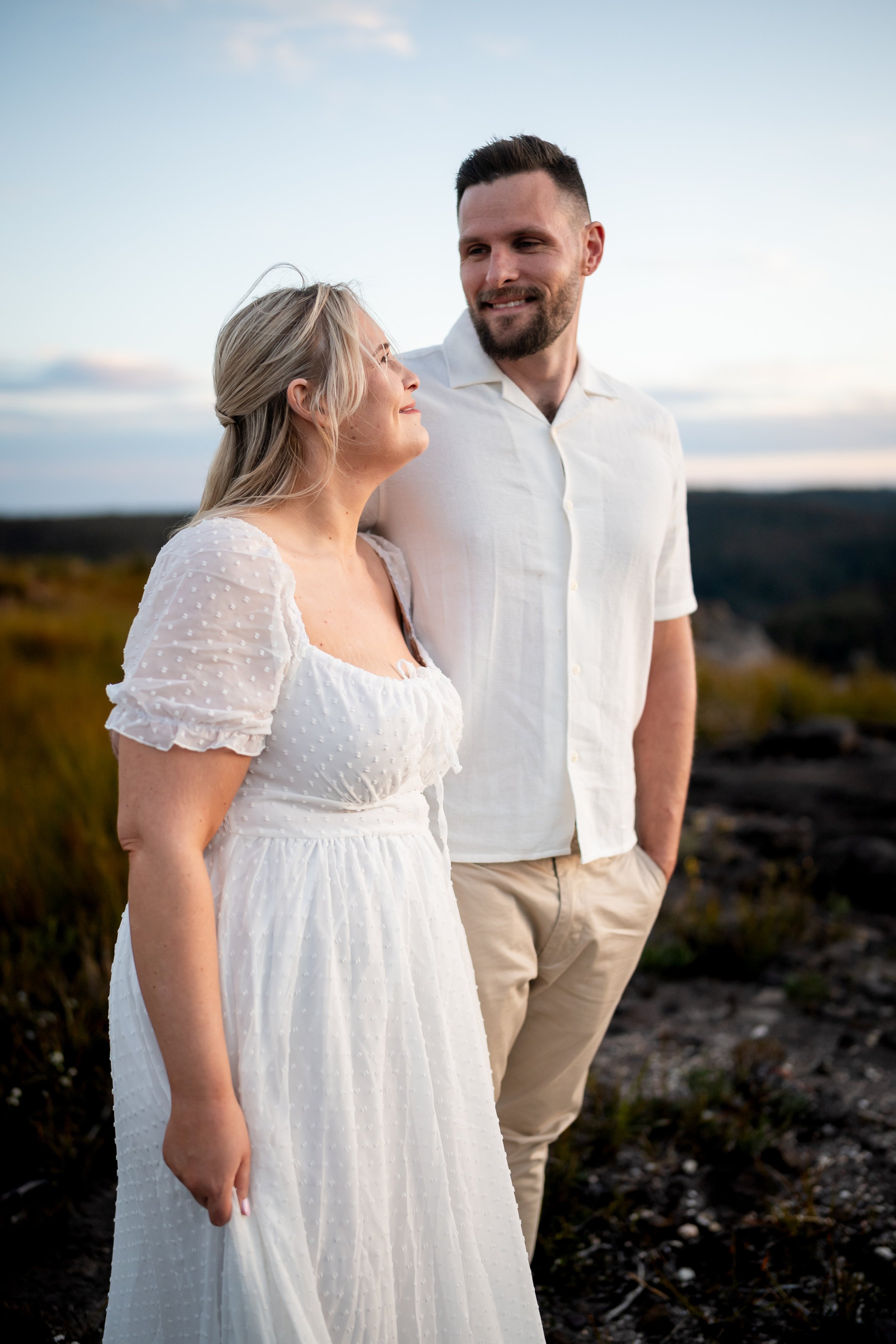 A couple standing outdoors in a natural setting during sunset, with the woman in a white dress and the man in a white shirt and beige pants, looking at each other lovingly.