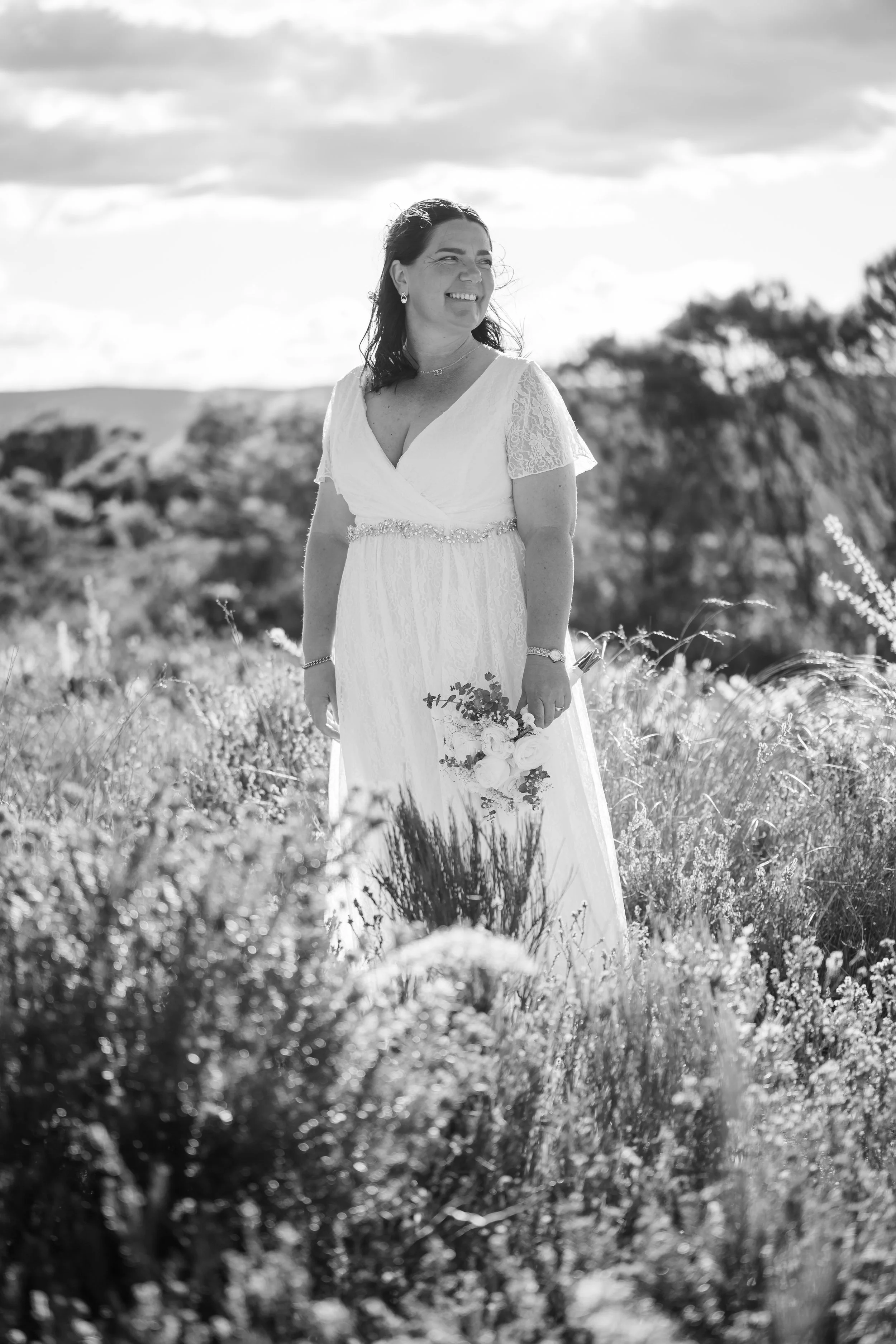 A smiling woman in a white wedding dress holding a bouquet of flowers stands in an outdoor field with tall grass and plants, with trees and a cloudy sky in the background.