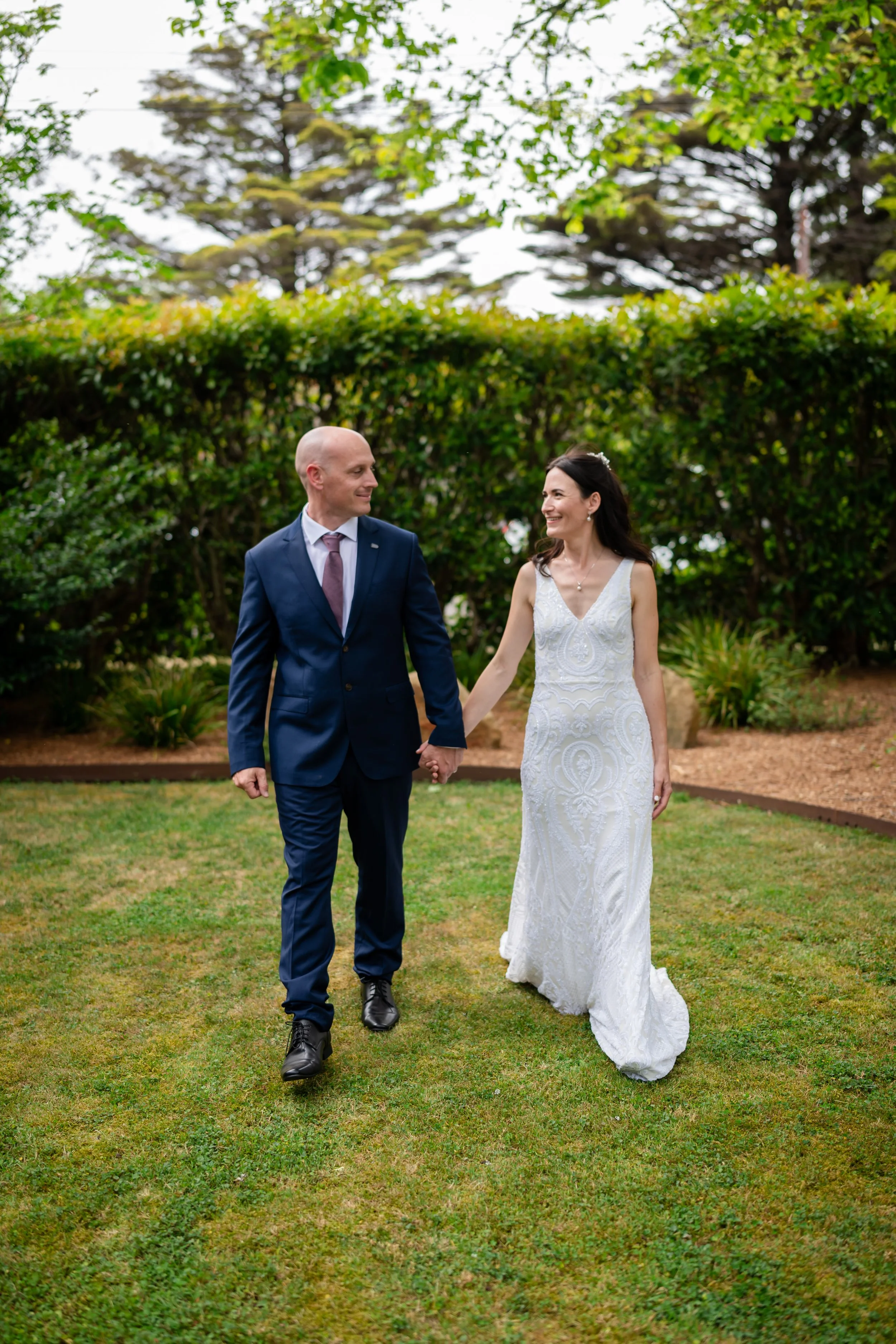 A bride and groom holding hands and walking on grass in a garden.