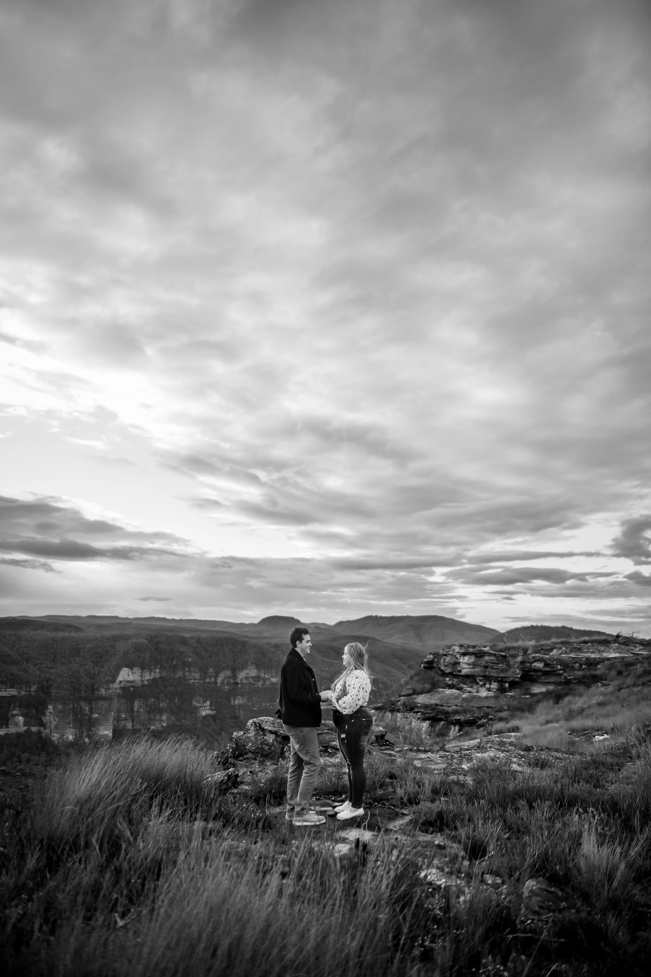 A couple holding hands and facing each other on a rocky ledge overlooking a canyon landscape during cloudy weather in black and white.