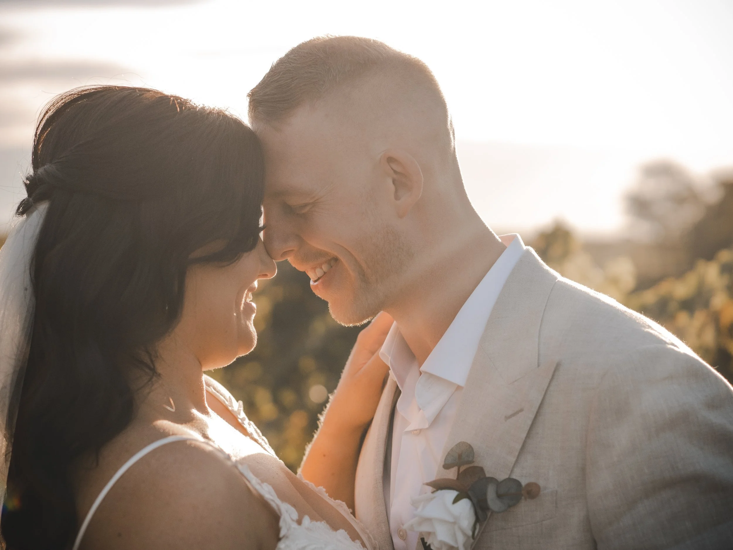 A happy couple, the bride and groom, with their foreheads touching and smiling, outdoors on their wedding day during sunset.