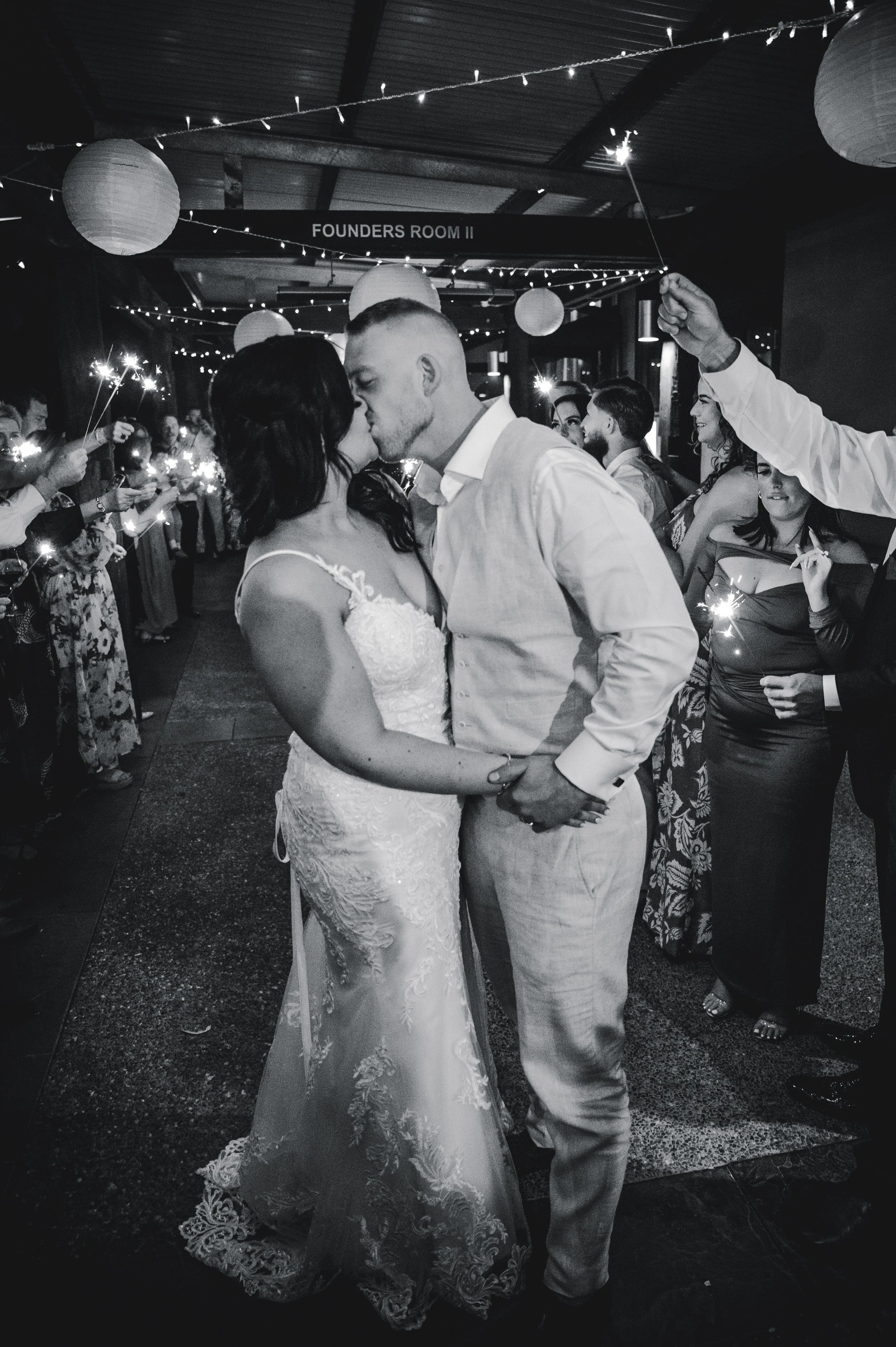 A black and white photo of a wedding reception with a bride and groom sharing a kiss in the center, surrounded by guests celebrating and holding sparklers.