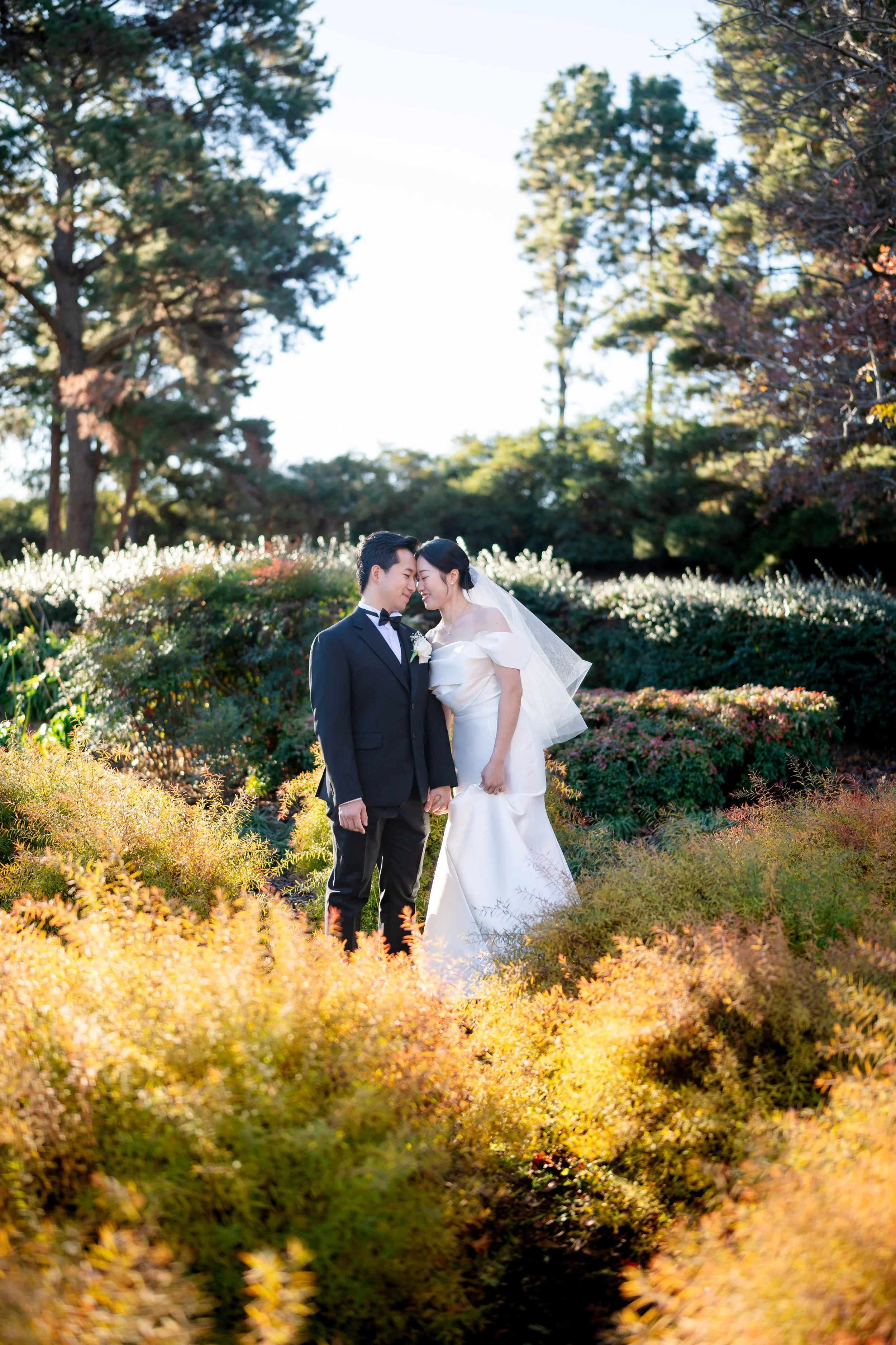A bride and groom standing close together outdoors in a garden, holding hands and touching foreheads, surrounded by colorful autumn foliage and bright sunlight.