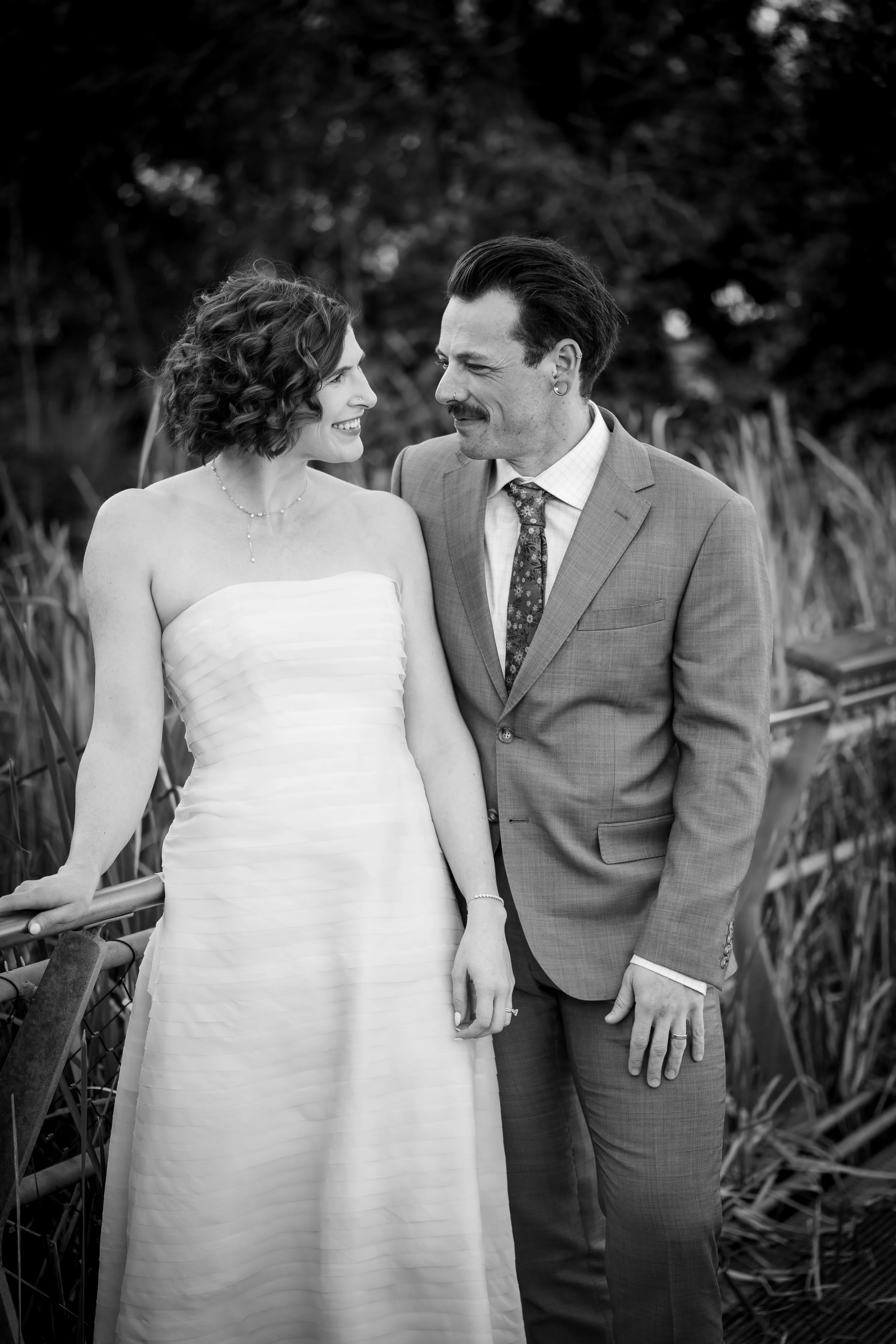 Black and white photo of a couple in wedding attire standing outdoors, smiling at each other. The woman is wearing a strapless wedding dress, and the man is in a suit with a patterned tie. They are holding hands near a railing with tall grass and tre