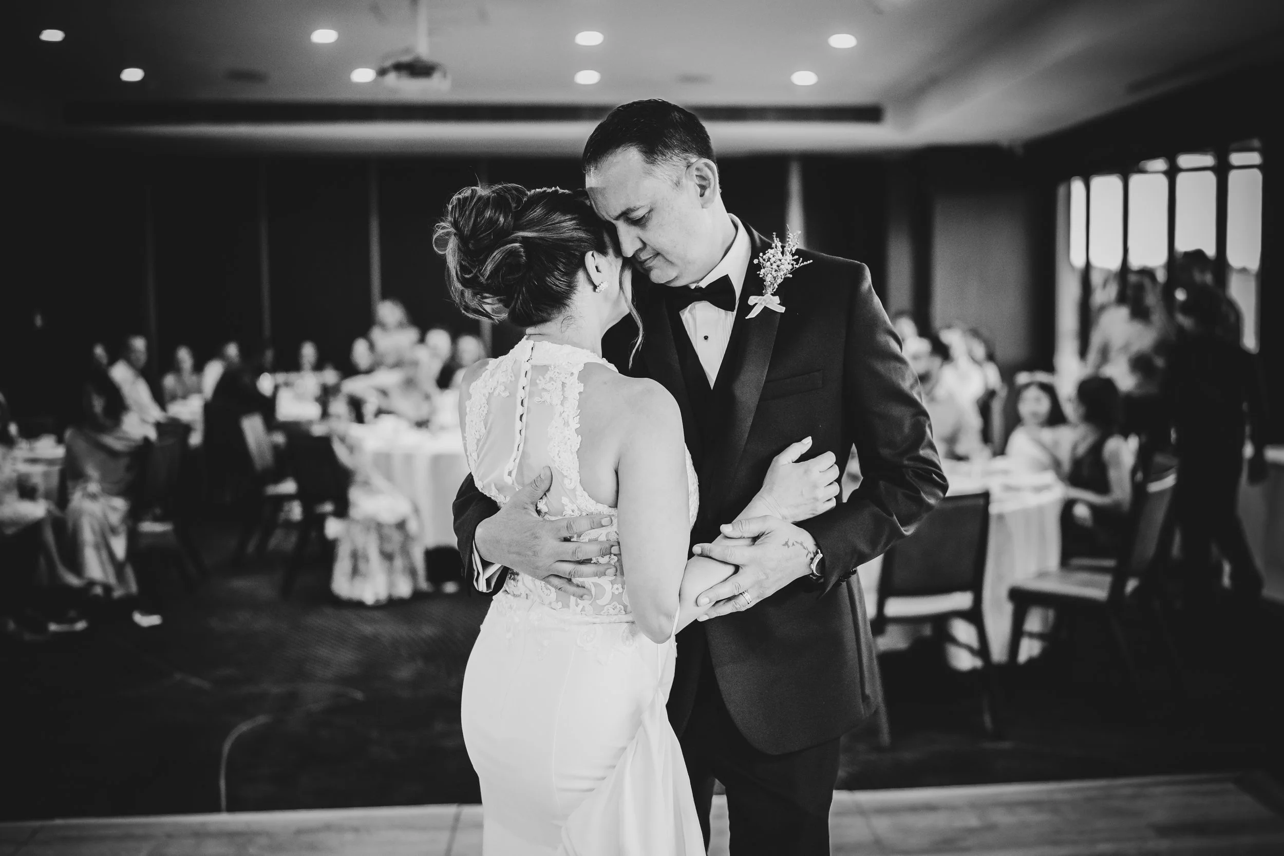 A black-and-white photo of a bride and groom sharing a dance at their wedding reception, surrounded by guests seated at tables in the background.