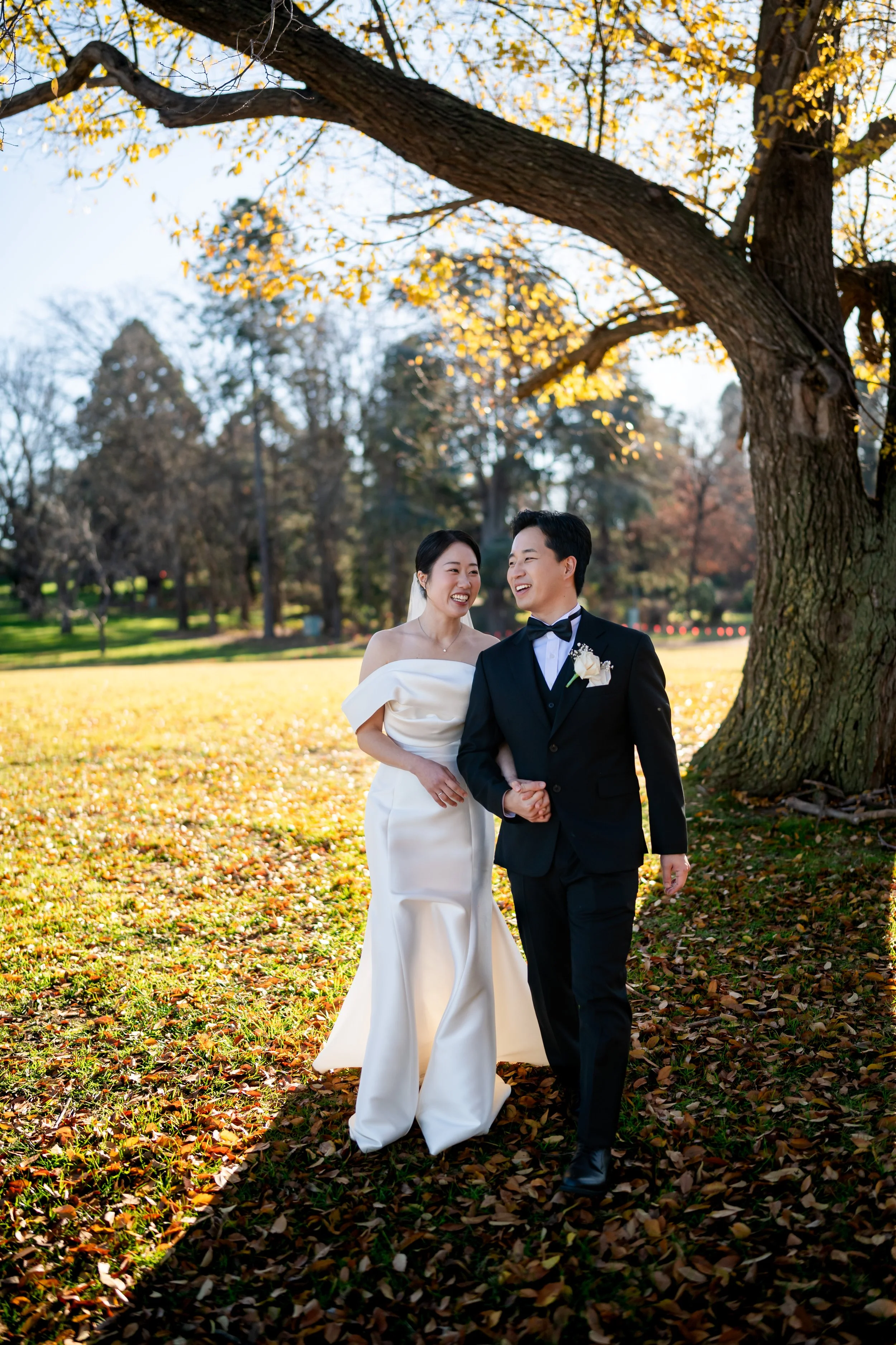 A bride and groom walking hand in hand outdoors on a sunny autumn day, surrounded by fallen leaves and trees with yellow and orange foliage.