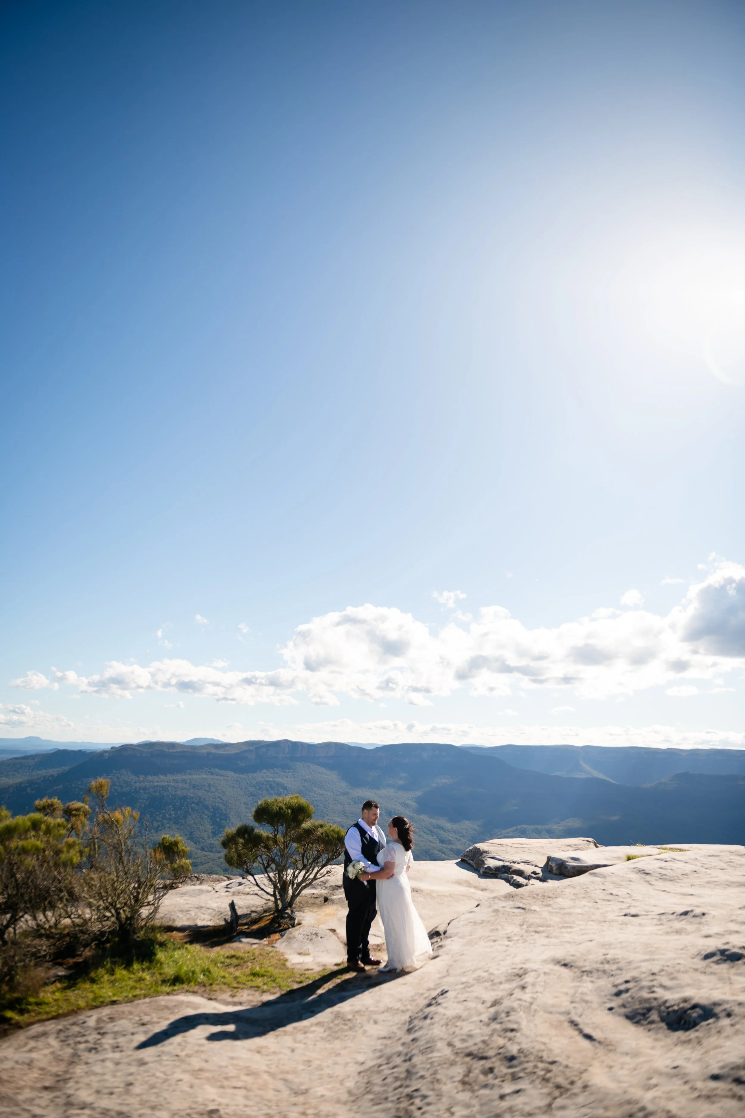 A bride and groom standing on a rocky ledge with mountain views in the background, during a sunny day with a partly cloudy sky.