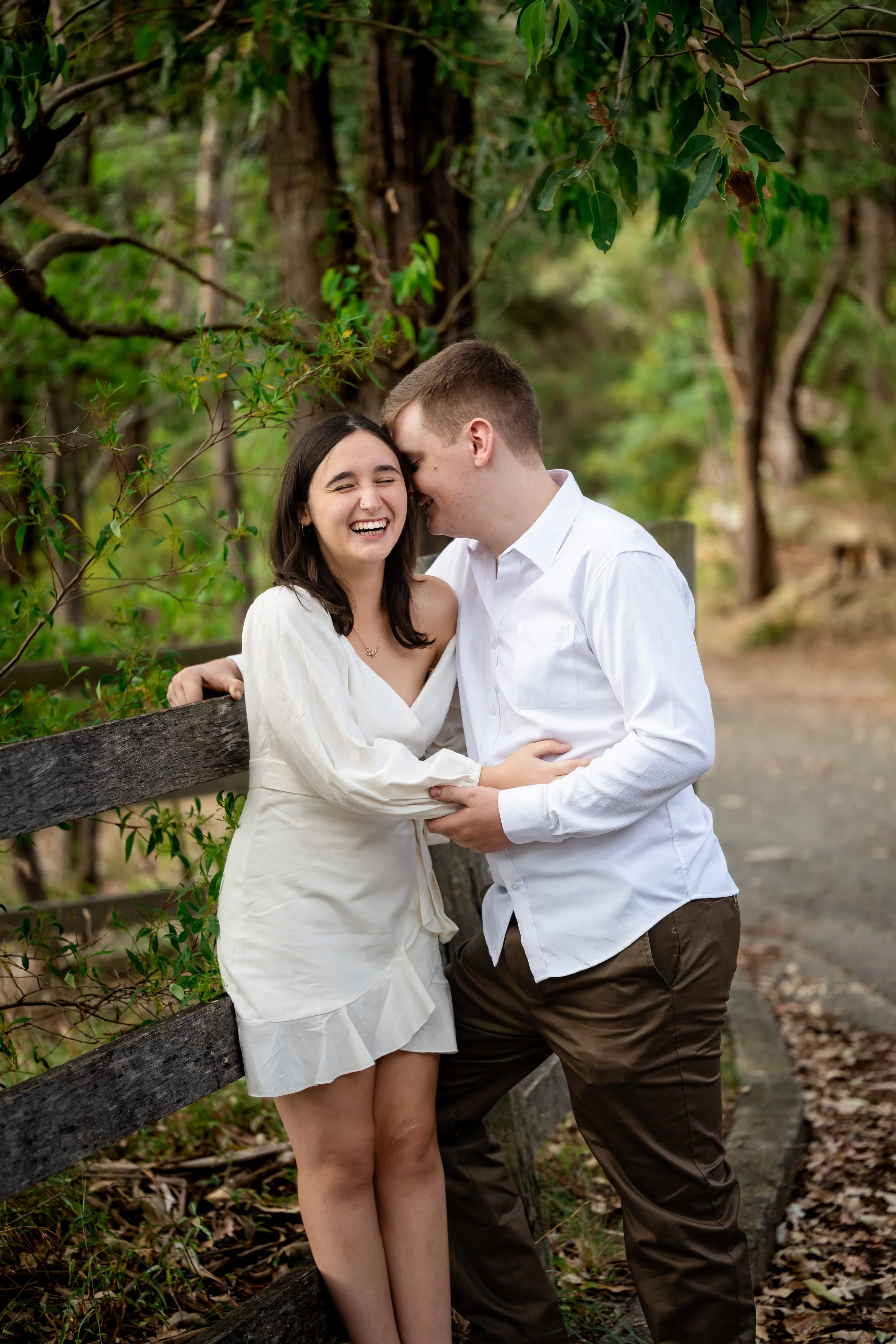 A young couple sharing a joyful moment outdoors, standing beside a wooden fence surrounded by green trees, with the woman smiling and the man leaning in close.