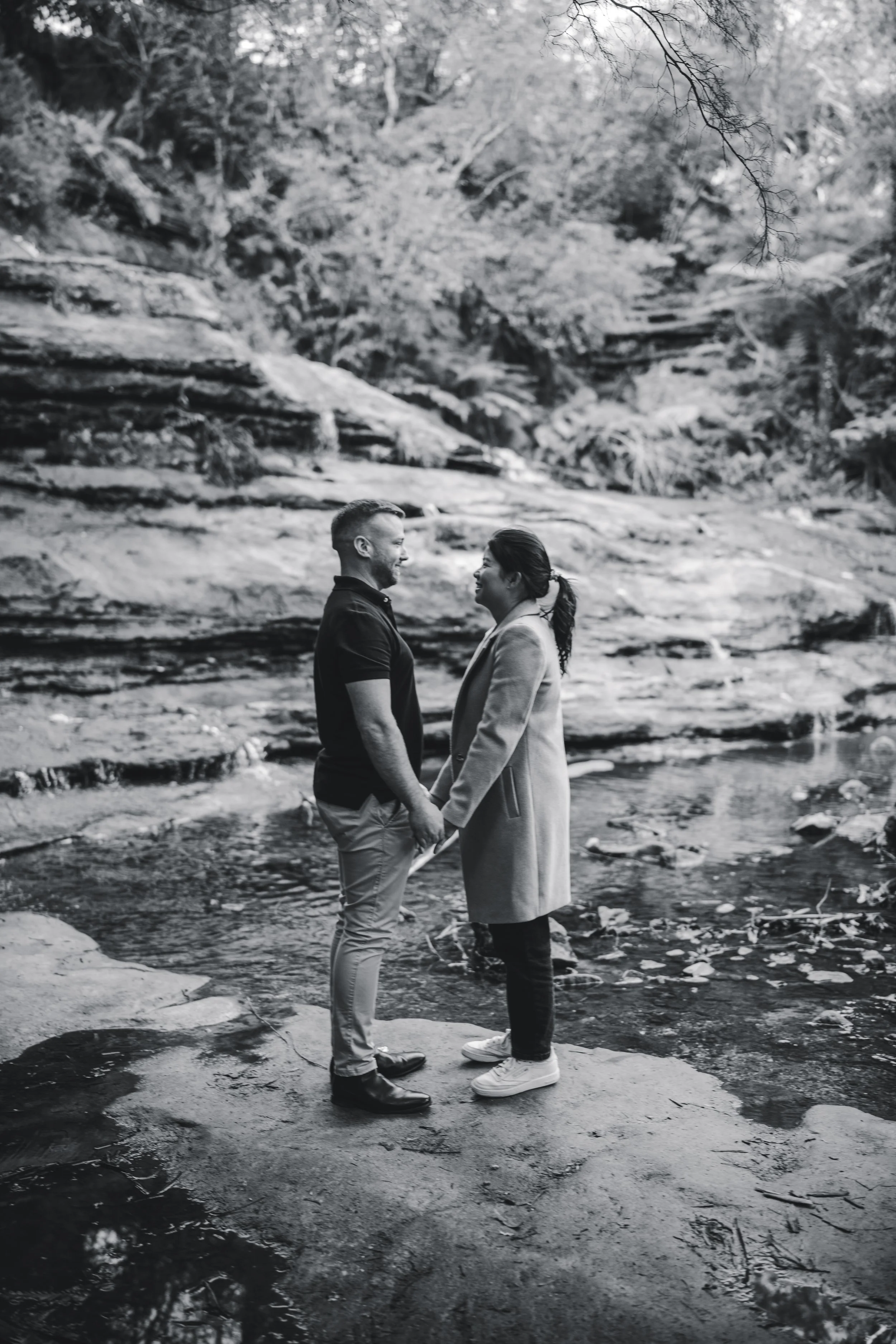 A couple standing hand in hand on a rock in a wooded creek, smiling at each other.